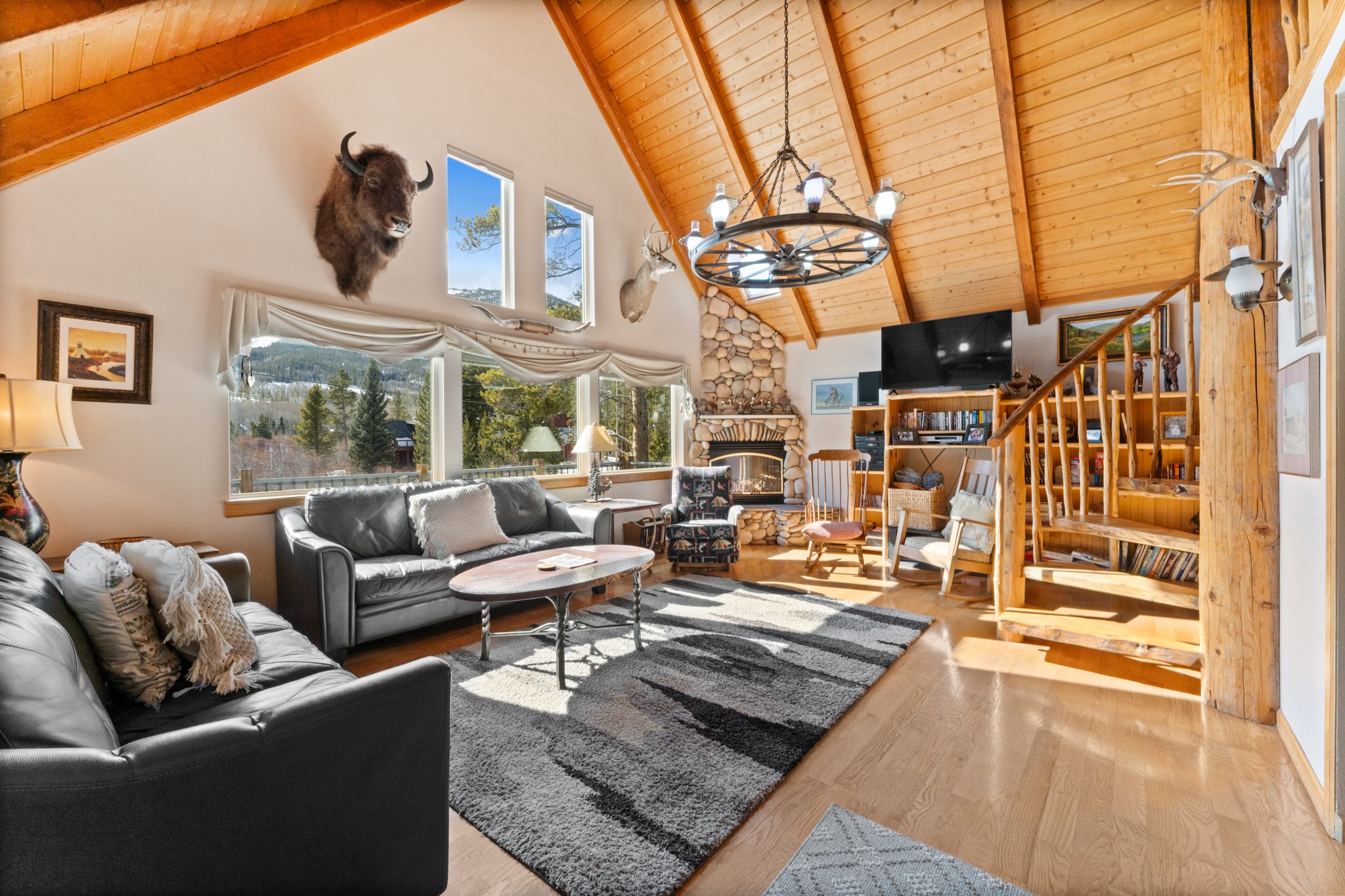 Living room with wooden vaulted ceiling, large windows showing mountain landscape, and decor including animal mounts and a stone fireplace. Contains leather sofas, an area rug, wooden chairs, and a staircase.