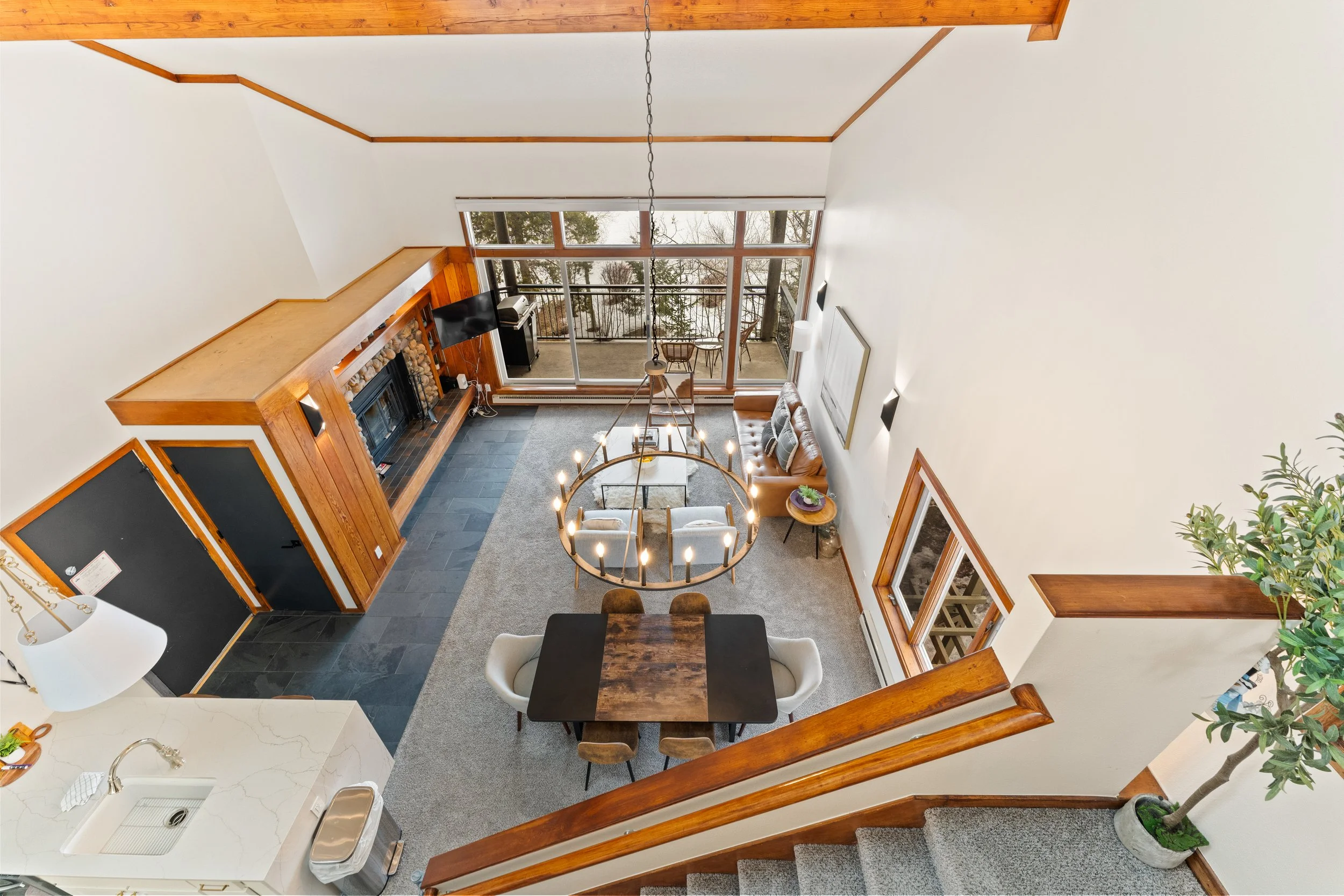 Interior view of a living room and dining area with large glass doors leading to a balcony. The room features a fireplace with a wood mantel, a dining table with six chairs, a leather sofa, a chandelier, and outdoor furniture on the balcony. The spac