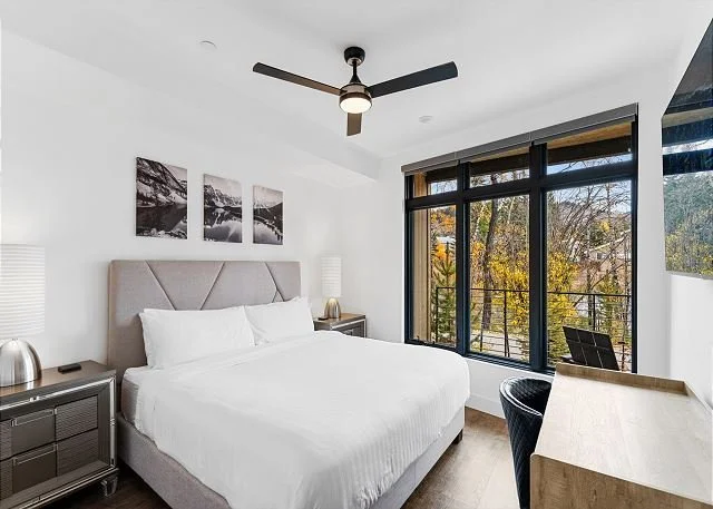 A modern bedroom with a large bed, nightstands with lamps, a ceiling fan, a sliding glass door, and artwork above the headboard.