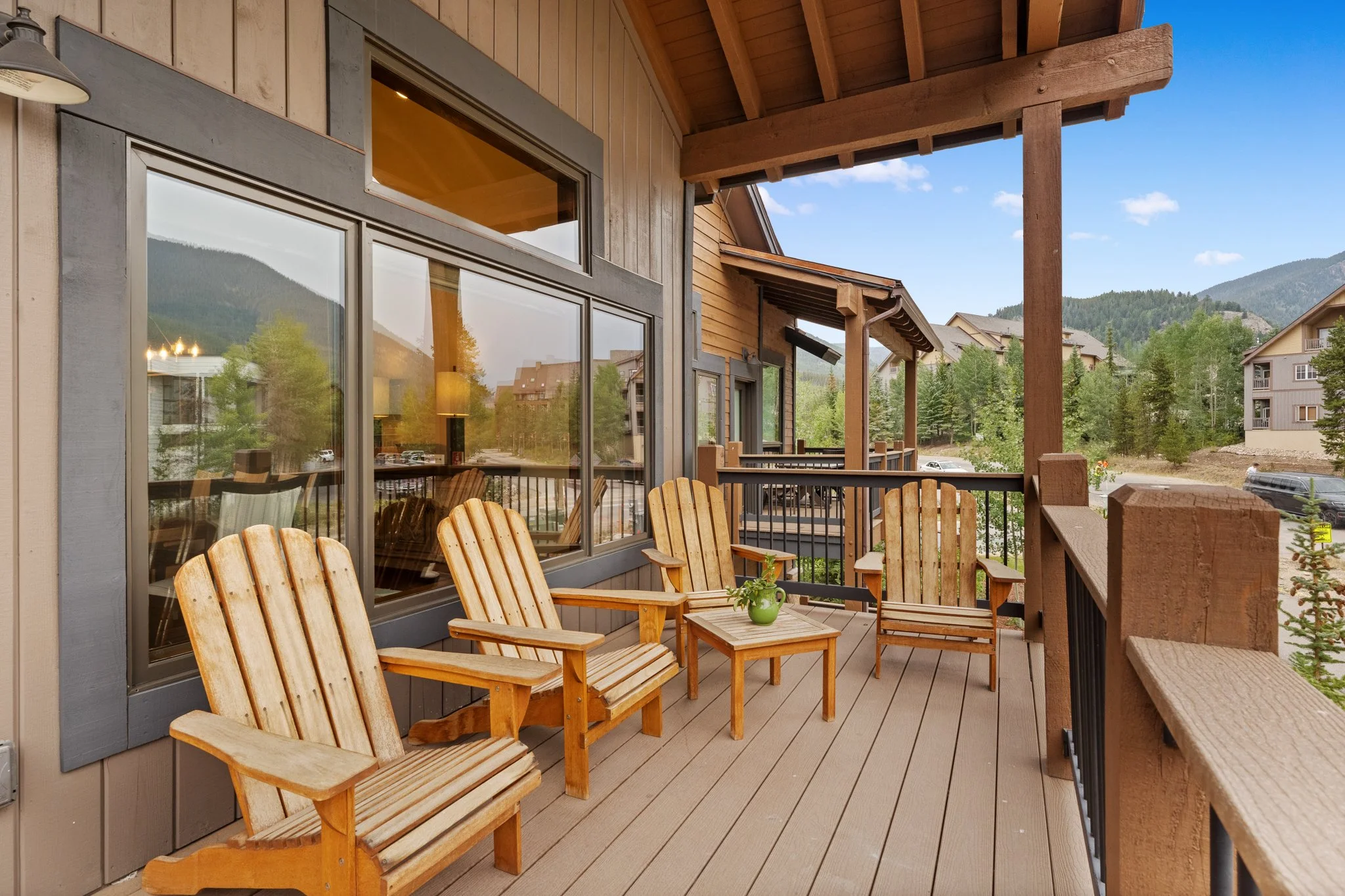 A wooden outdoor balcony with three Adirondack chairs and a small table with a potted plant, overlooking a mountain landscape with trees and neighboring buildings.