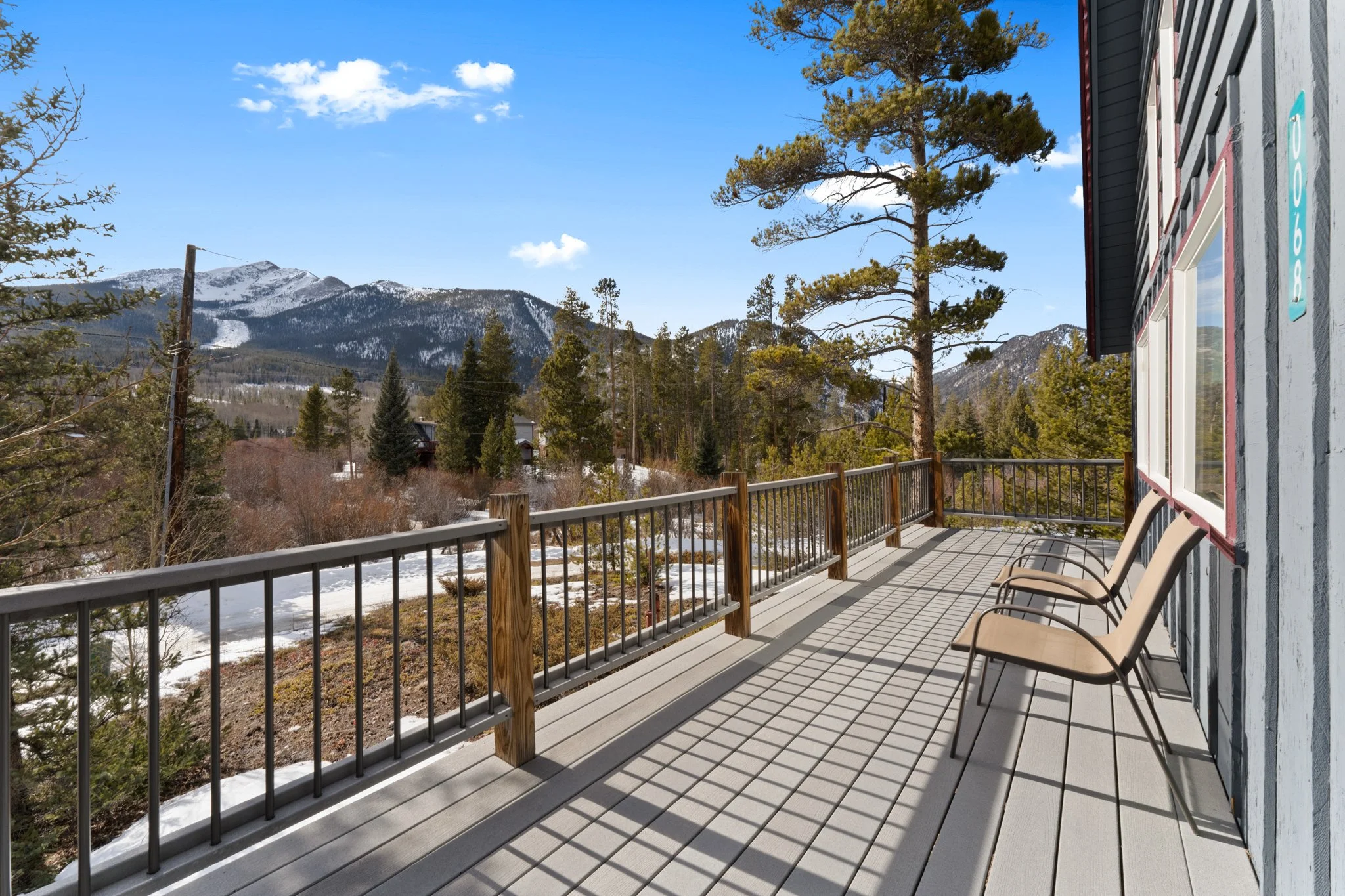 A wooden balcony with two beige chairs overlooking a mountain landscape with pine trees and some snow, under a blue sky with a few clouds.
