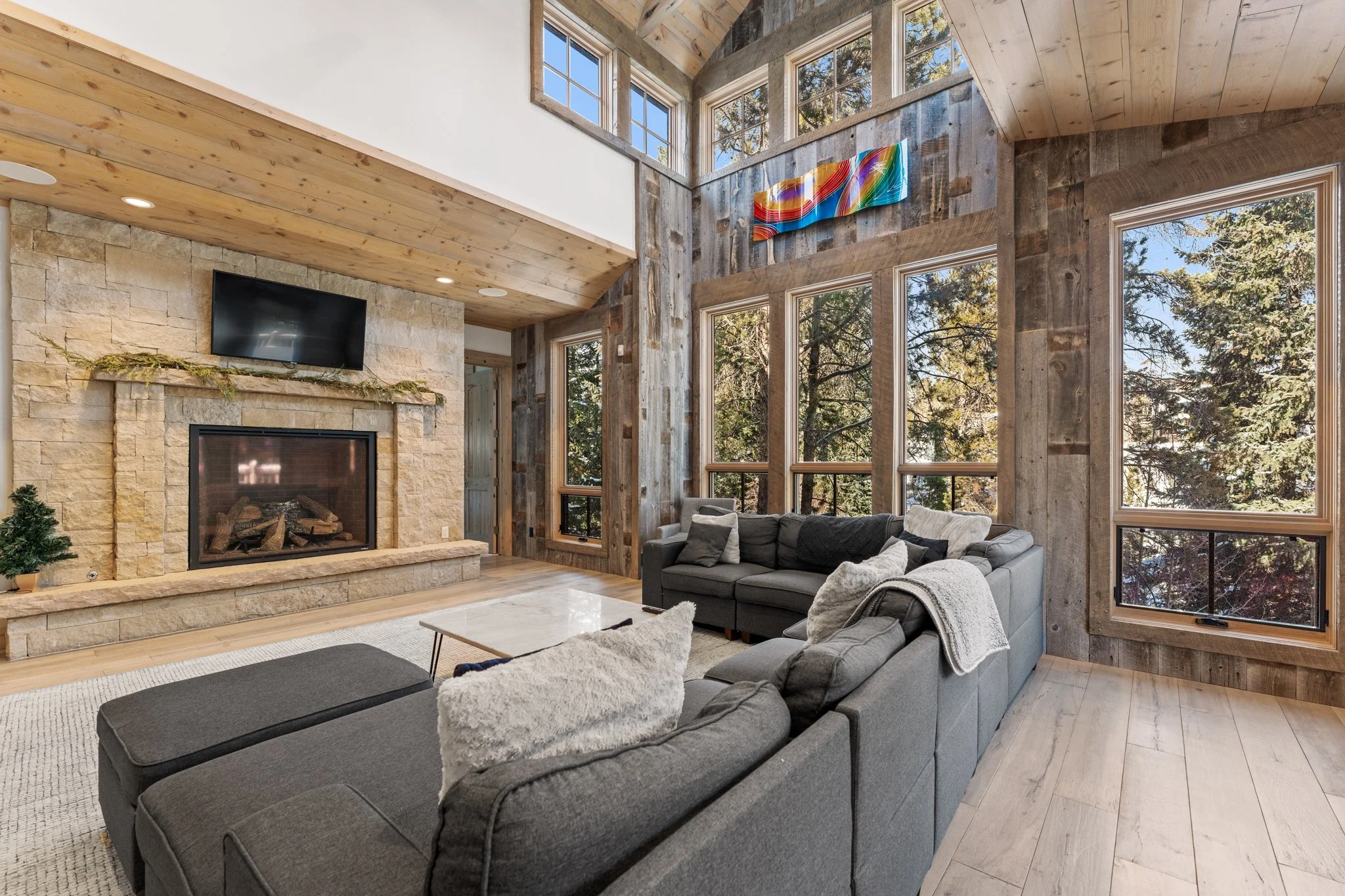Living room with large windows, stone fireplace, gray couches, wooden walls, and a wall-mounted TV.
