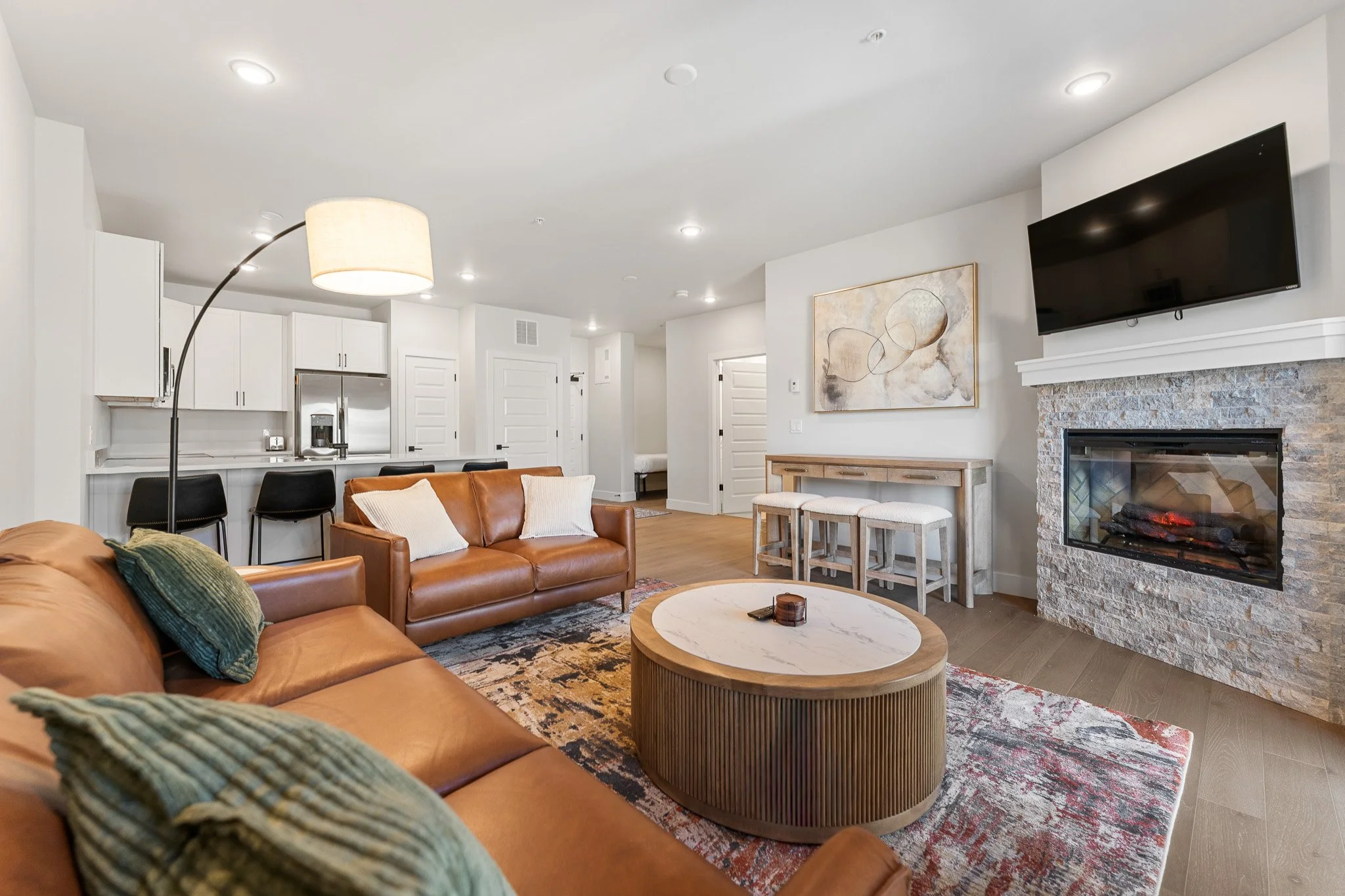 Living room with brown leather sofas, a round coffee table, a stone fireplace with a mounted TV, white walls, artwork, and a bar area with black chairs in the background.
