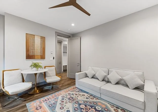 Living room with a white sofa, three pillows, a round table with a plant, two chairs, a colorful area rug, a wooden ceiling fan, and an open door leading to another room.