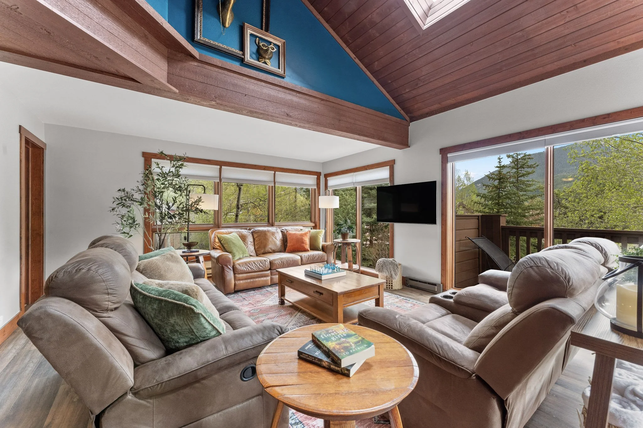 A cozy living room with large windows showing green trees and mountains outside. It has a vaulted ceiling with wood paneling, beige and brown sofas, a wooden coffee table, and a flat-screen TV on the wall.