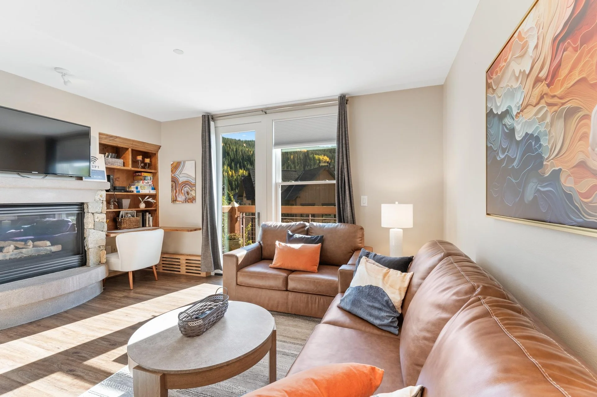Living room with leather sofas, a stone fireplace, a wooden shelf with books, a flat-screen TV, a window showing a mountain view, and abstract wall art.