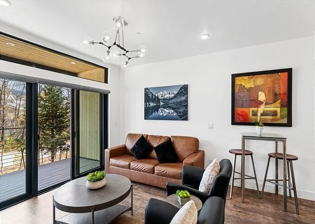 Living room with sofa, chairs, coffee table, artworks on wall, and sliding glass doors leading to balcony with trees outside.