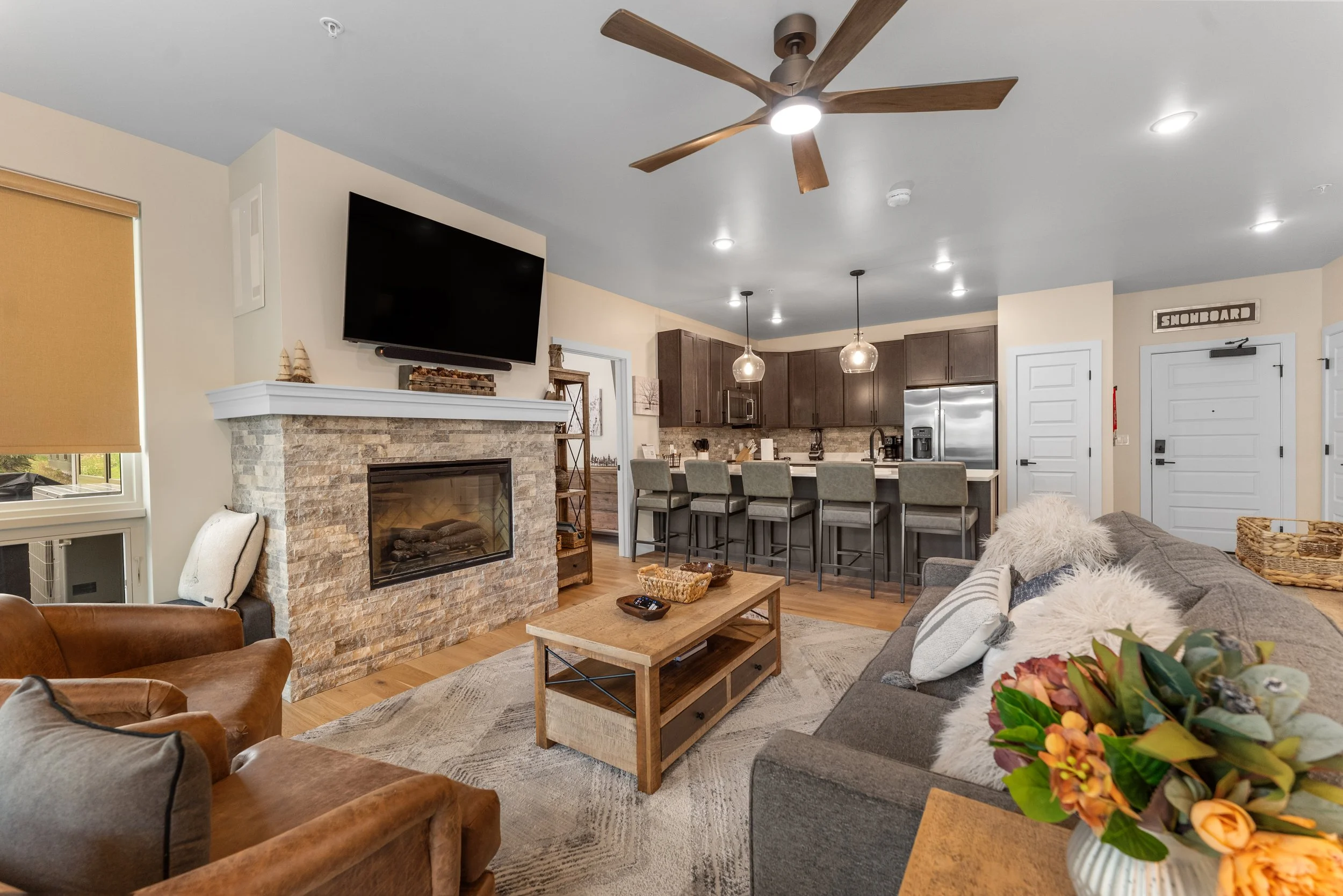 Living room with a stone fireplace topped with a TV, a ceiling fan, a gray sofa with fluffy pillows, a coffee table, and a kitchen with a breakfast bar and pendant lights.