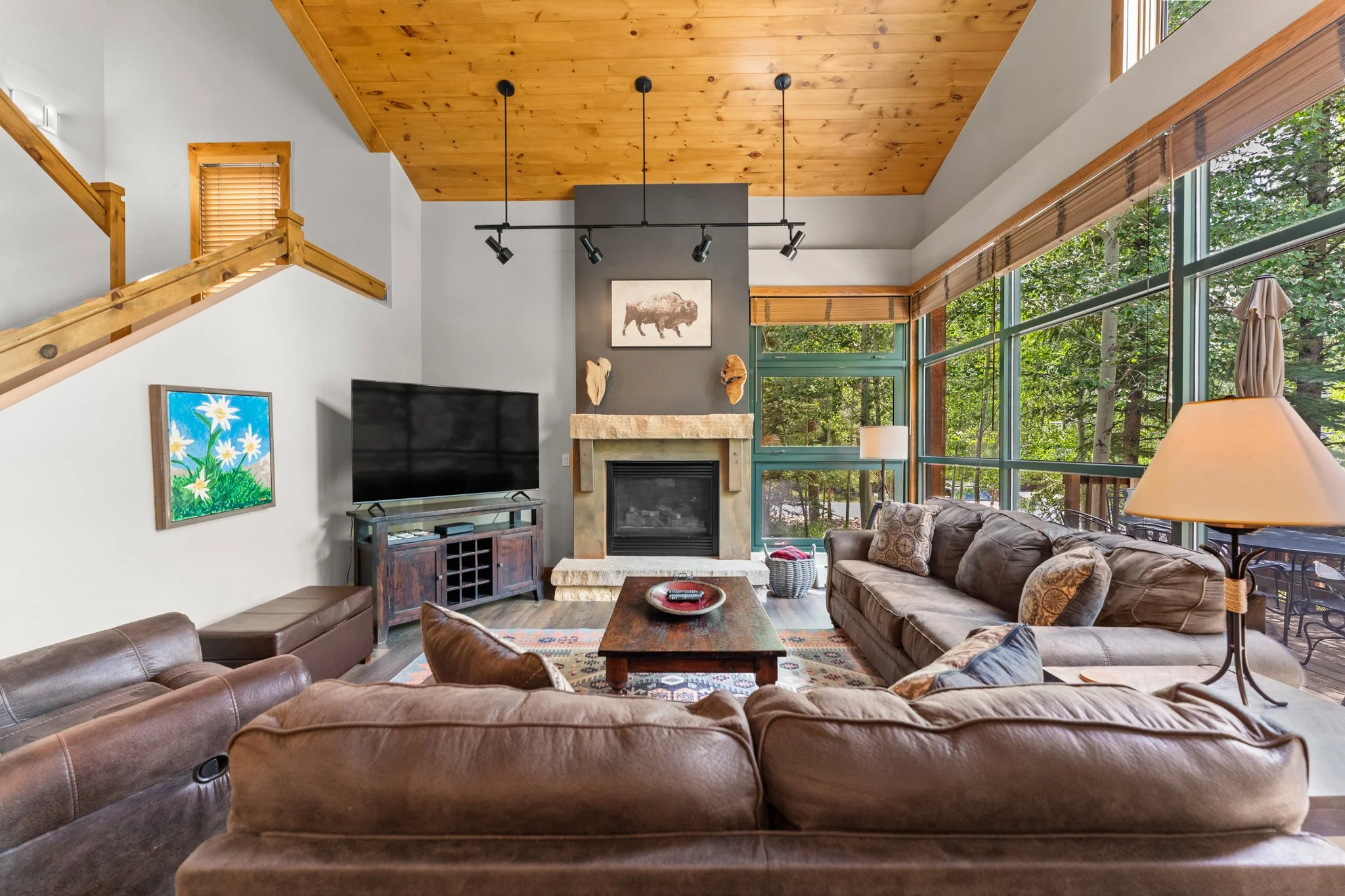 Living room with large windows, brown leather sofas, a wooden coffee table, a stone fireplace with artwork, and a wood-paneled ceiling.
