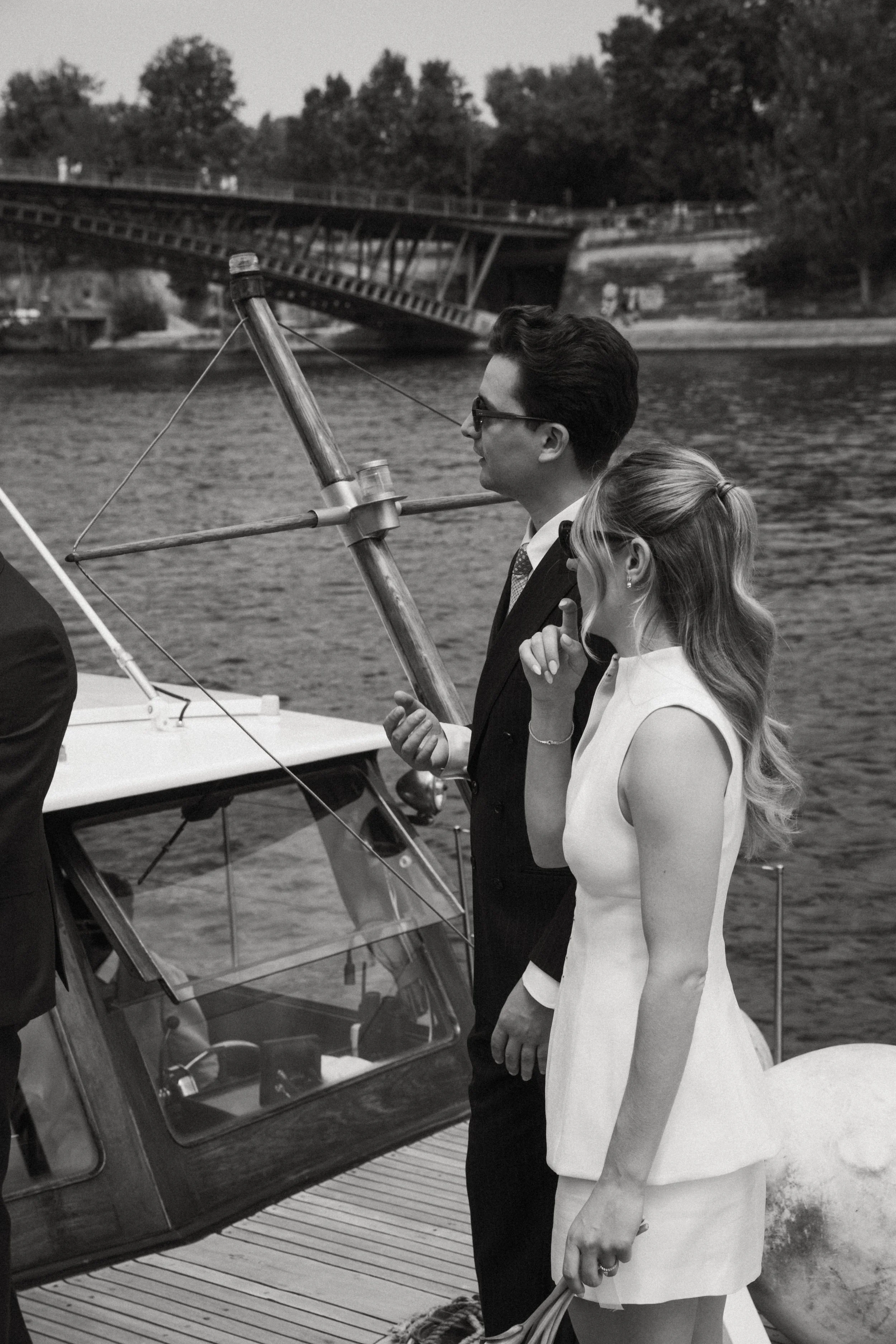 Un homme et une femme habillés élégamment, debout au bord de l'eau, près d'un bateau, lors d'une occasion formelle. La femme porte une robe blanche et le homme un costume noir.