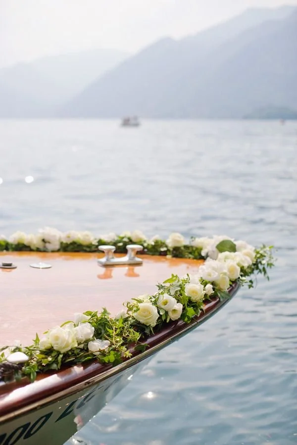 Proue d'un bateau décorée de fleurs blanches et feuillage, sur un lac avec montagnes en arrière-plan.
