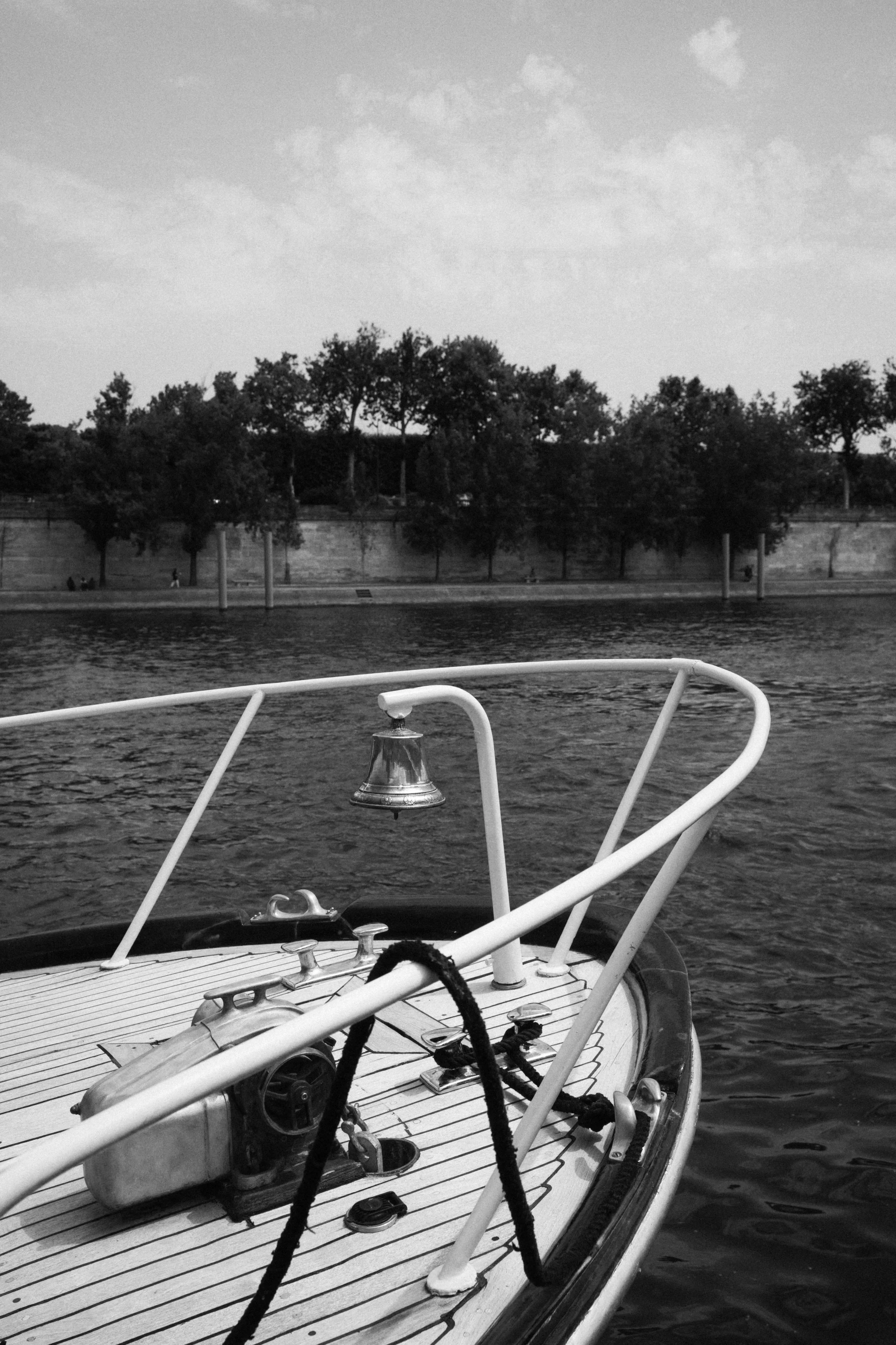 Vue du pont d'un bateau avec des cordages et un moteur, regardant vers une rive avec des arbres et un mur, contre un ciel nuageux.
