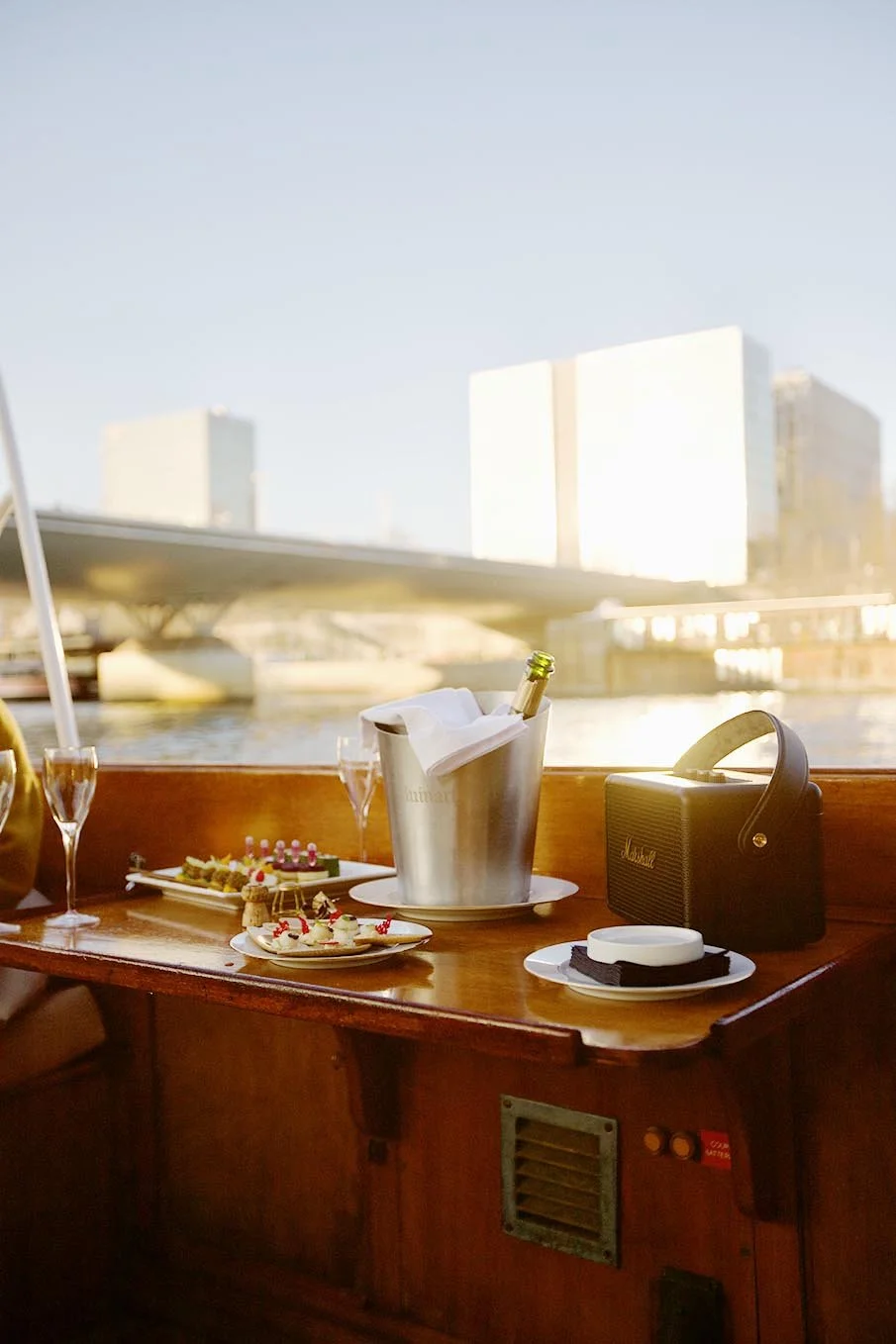 Table sur un bateau avec des amuse-bouches, un seau à champagne, des verres, et une enceinte portable, vue sur un pont et bâtiments modernes en arrière-plan.