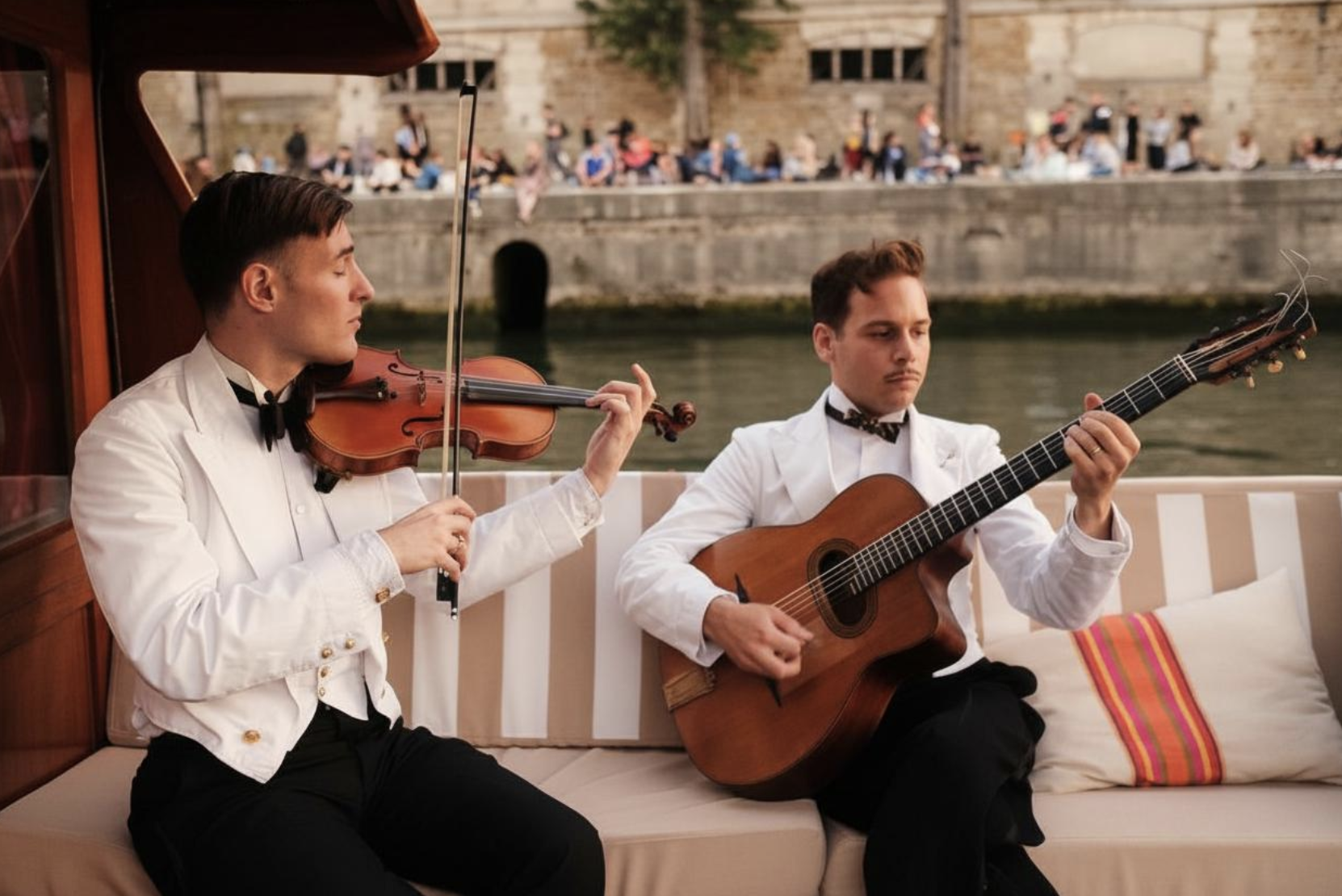 Deux musiciens habillés en veste blanche et nœud papillon noir jouent de la musique à bord d'une bateau près d'une rivière, avec un bâtiment en pierre en arrière-plan et un groupe de personnes assises sur la rive.