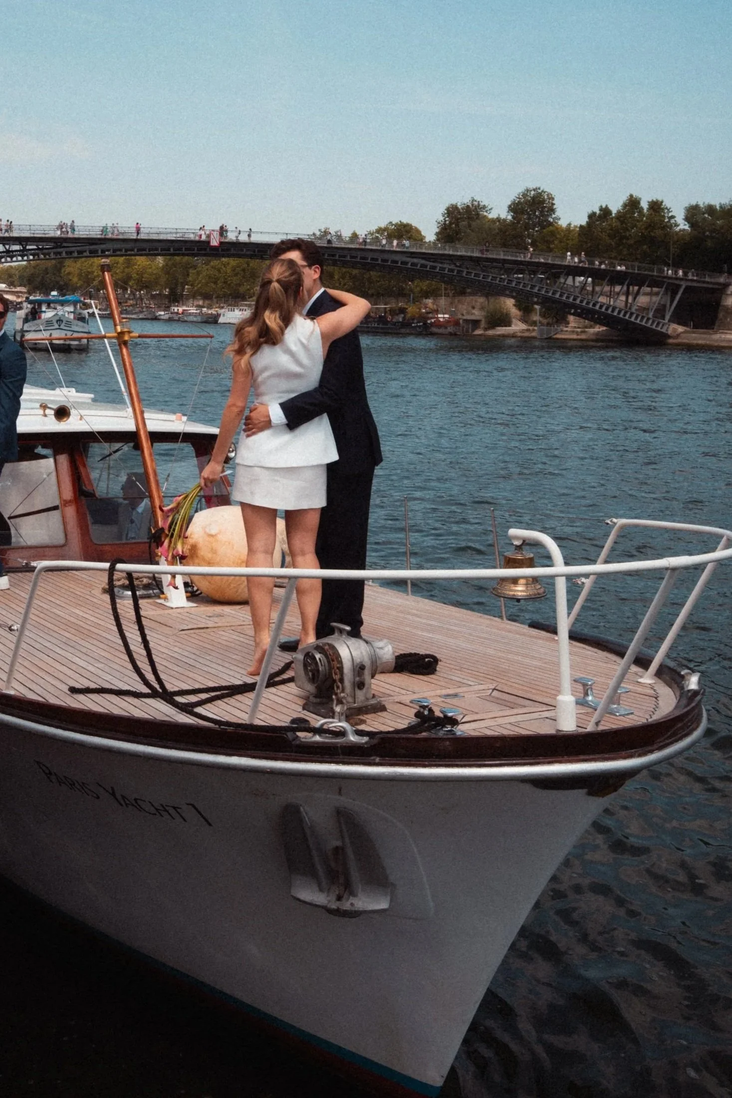 Un couple s'embrassant sur un bateau dans un port, avec un pont en arrière-plan et d'autres bateaux à quai.