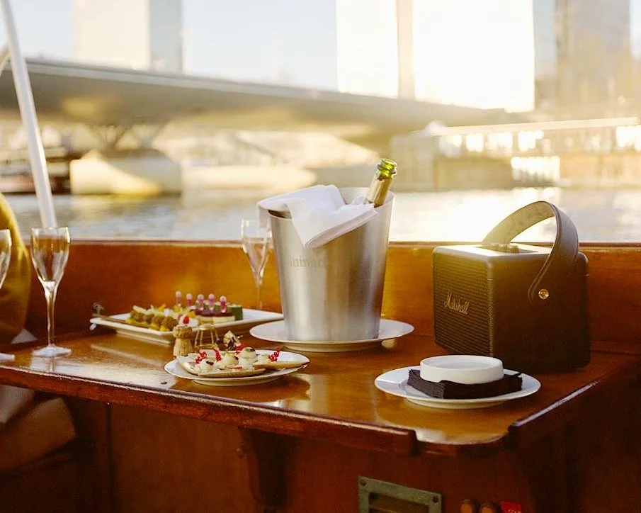 Vue de l'intérieur d'une cabine de bateau avec une table en bois, des desserts, une bouteille de champagne dans un seau à glace, un haut-parleur Marshall noir, et une vue floue d'un pont de bateau et d'une rivière en arrière-plan.