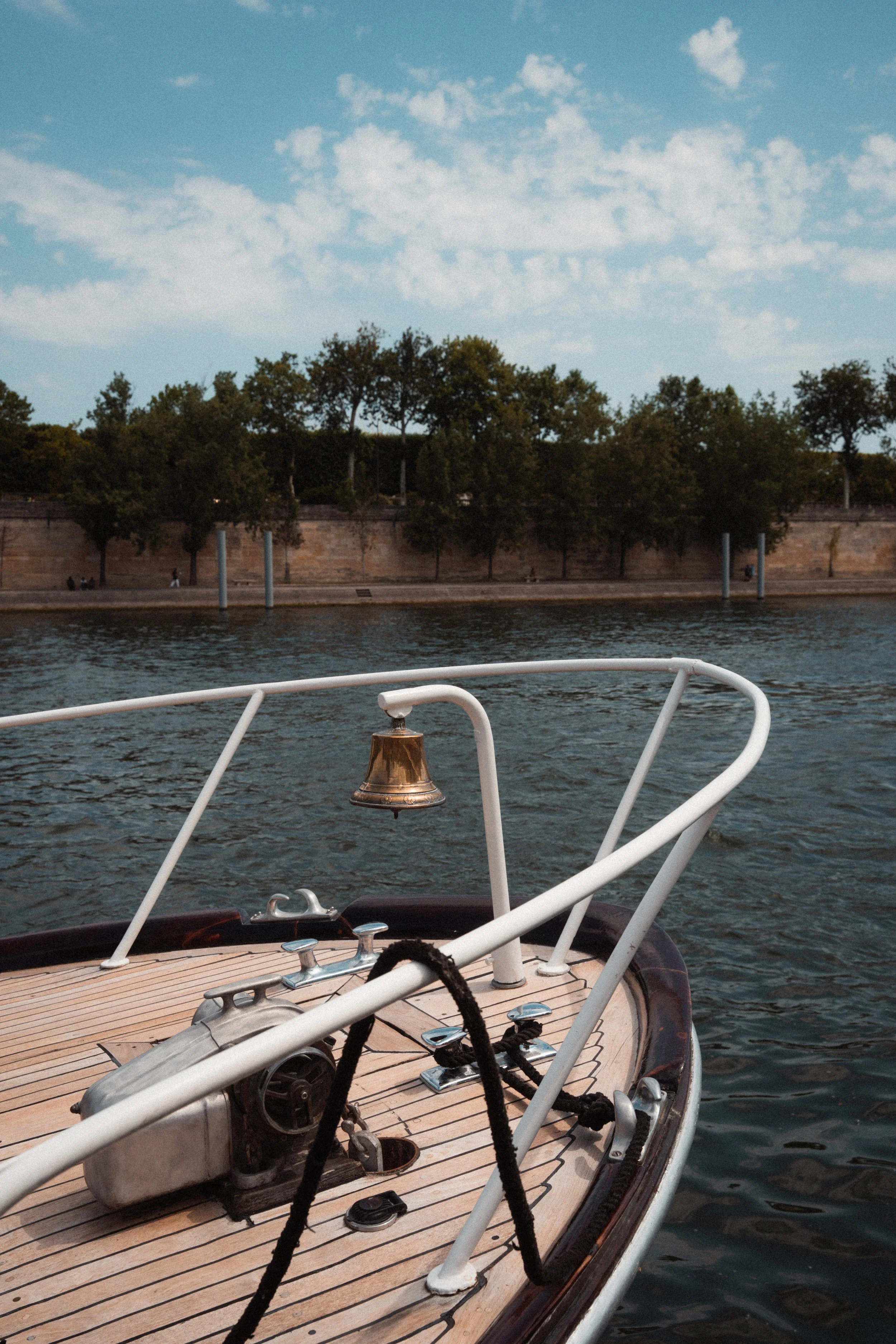 Vue du pont d'un bateau avec un moteur et des cordages, vue sur la rivière et la rive avec des arbres et un ciel partiellement nuageux.