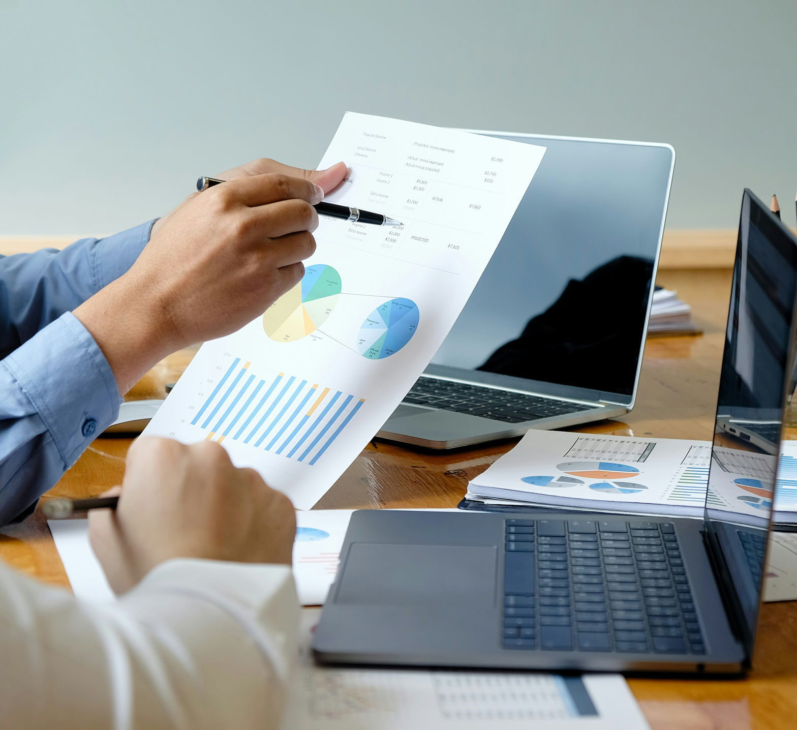 A person reviewing financial documents with graphs, charts, and pie charts, at a desk with a laptop, with another hand holding a pen.