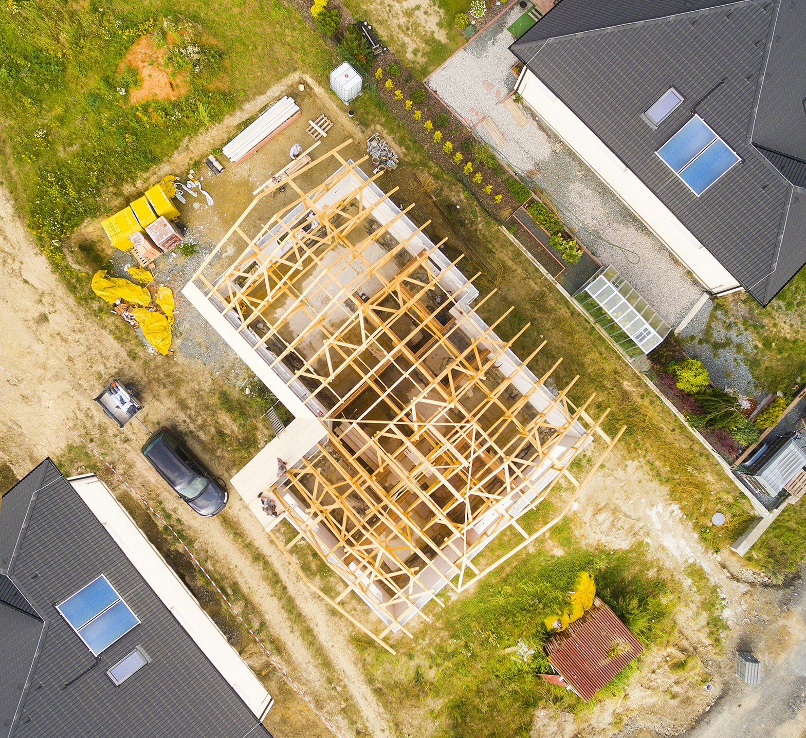 An aerial view of a house under construction with a wooden framework, surrounded by neighboring houses, lawns, vehicles, and construction materials.