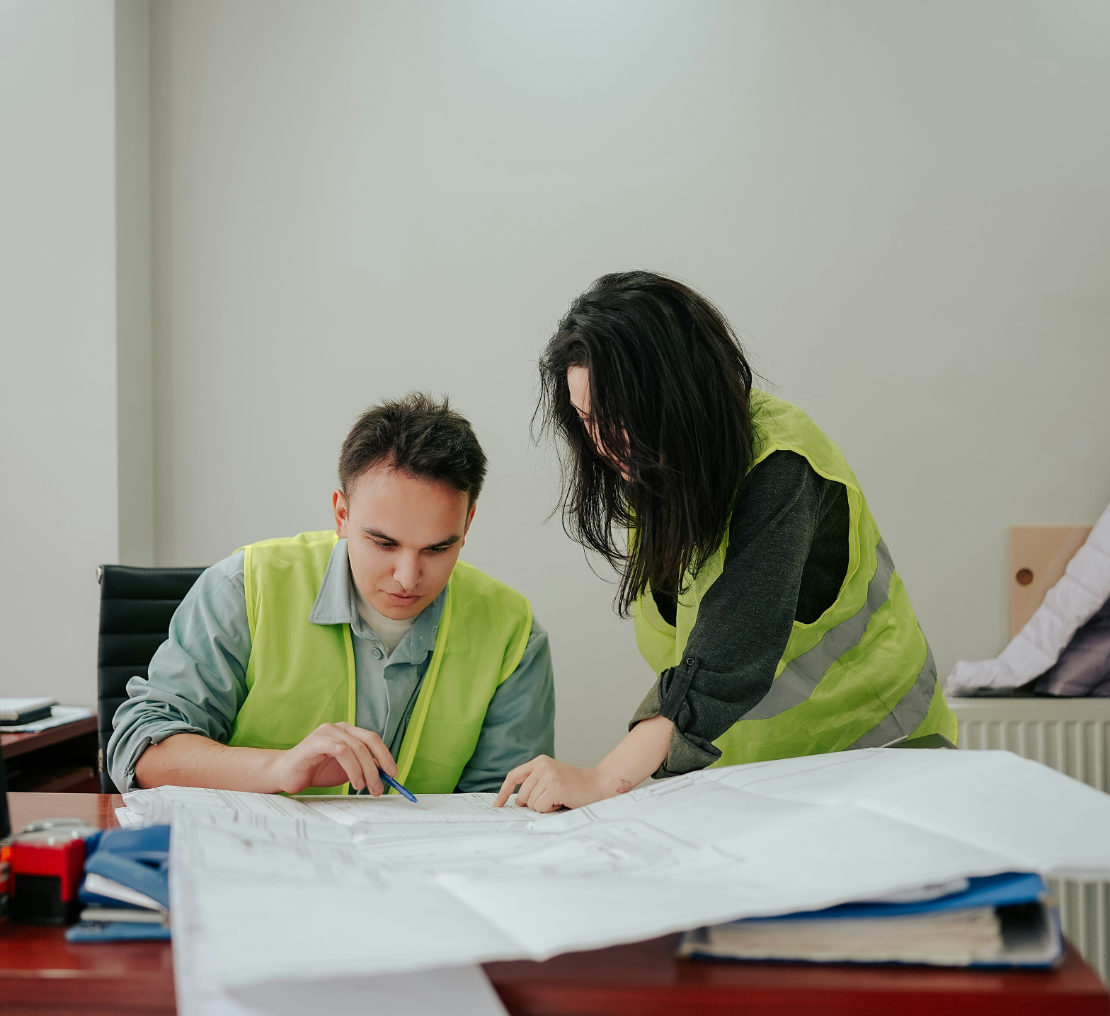 Two people wearing yellow safety vests working together on blueprints at a desk in an office or workspace.