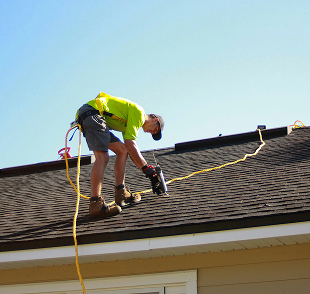 A person working on a roof wearing a bright yellow shirt, using a power drill, with a safety harness and rope for fall protection during daytime.
