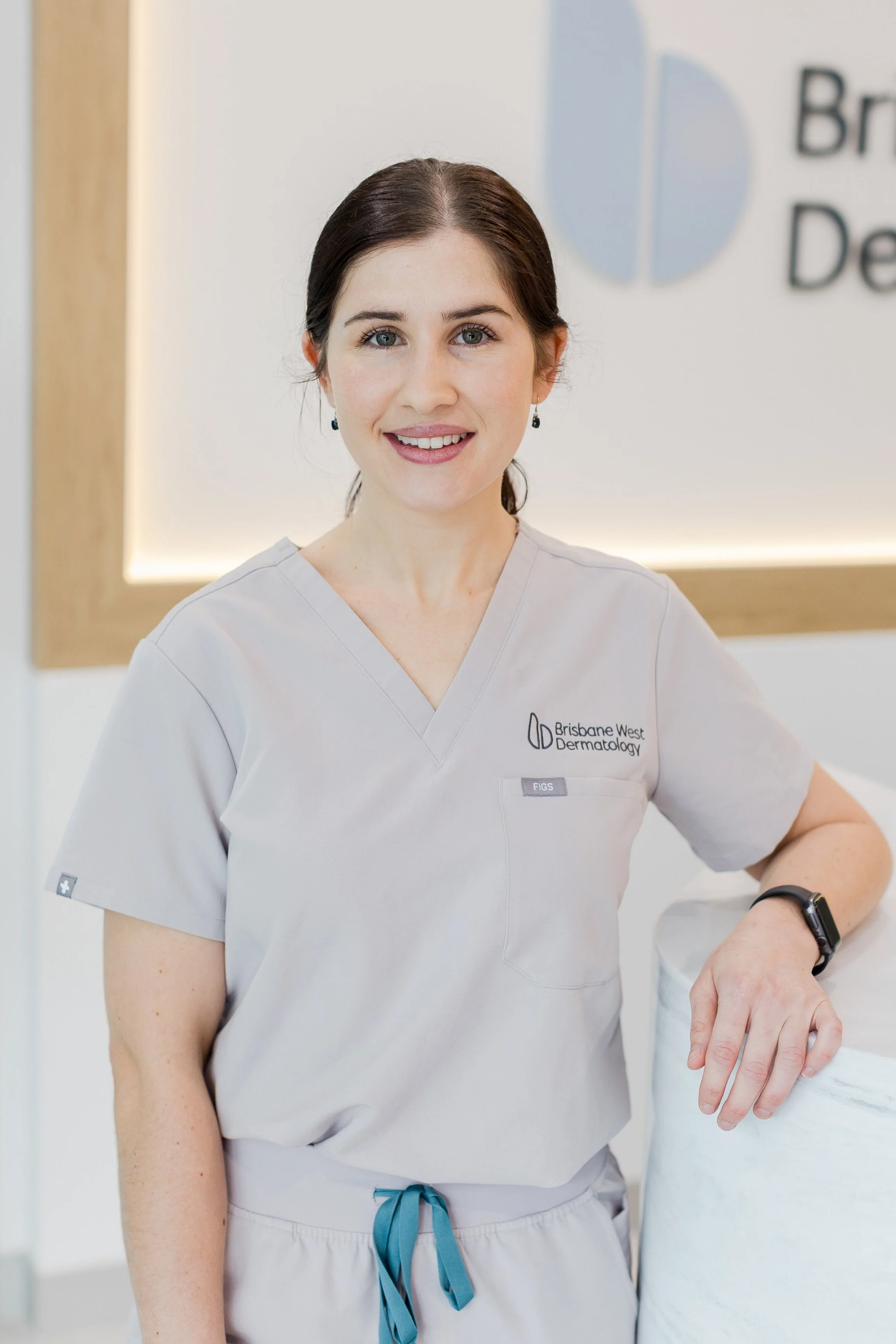 A smiling woman in grey scrubs with a Brisbane West Dermatology logo stands in a clinic, leaning her hand on a white marble counter.