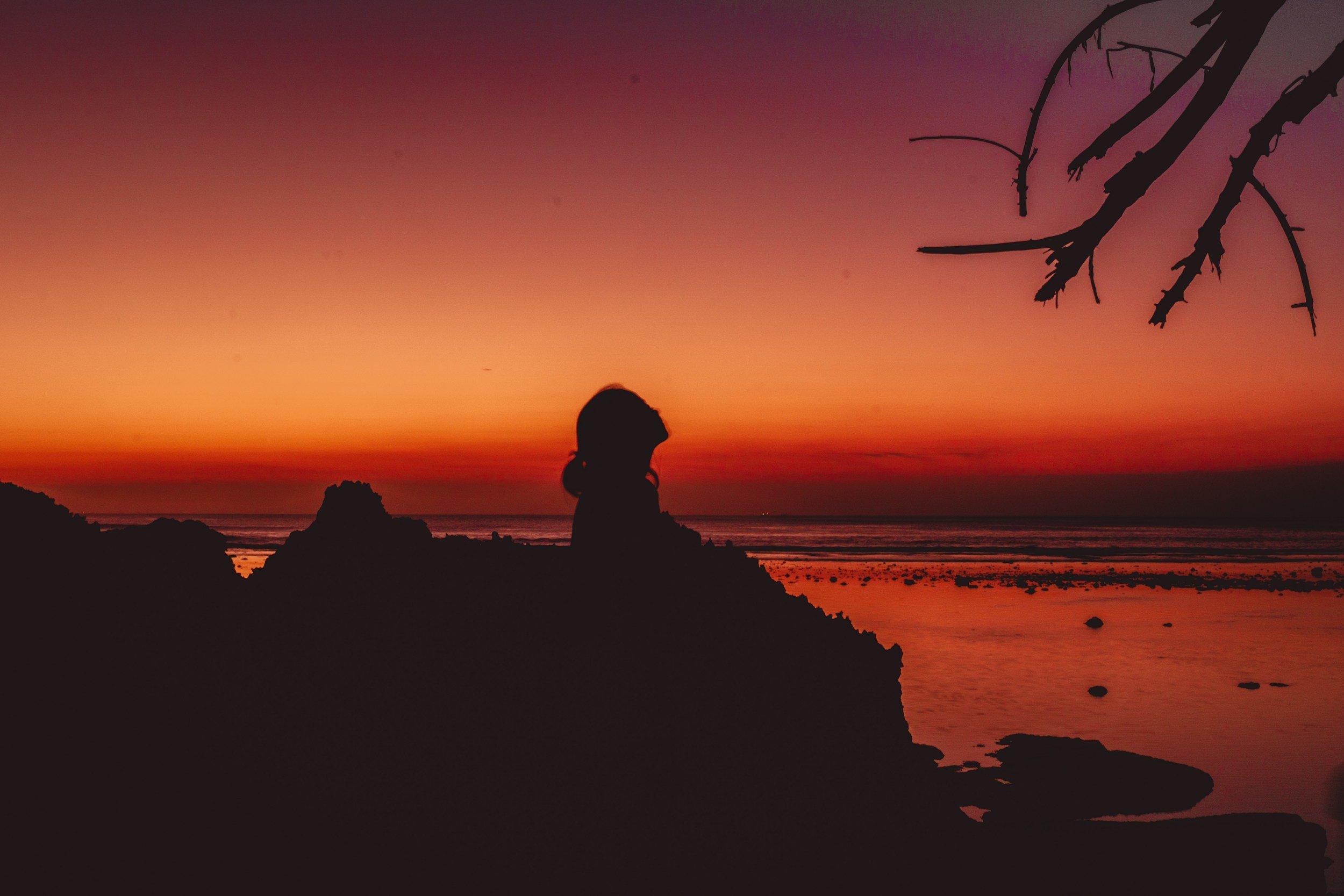 girl sitting on the beach looking at a bright orange and red sunset on the beach