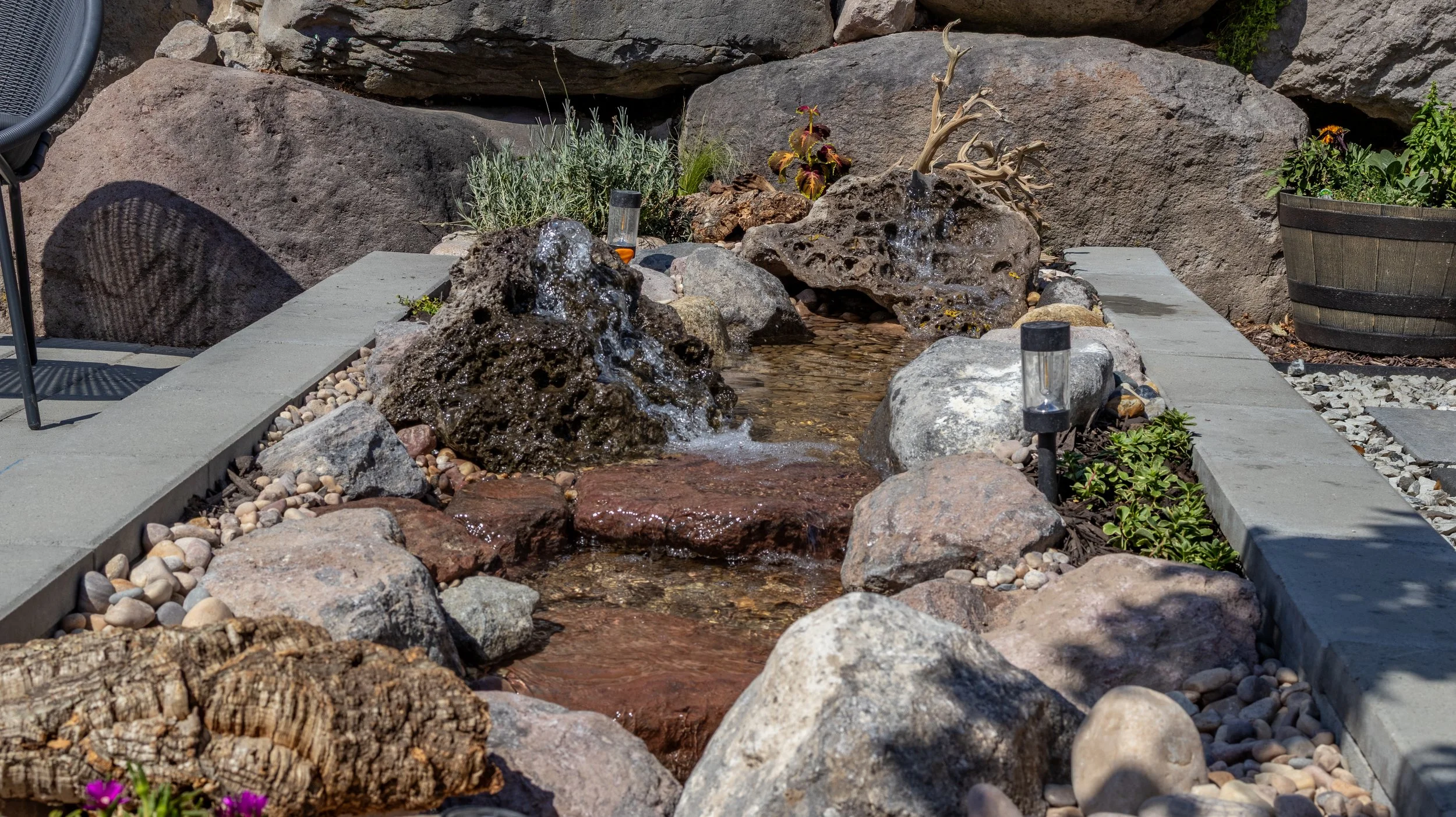 A small, decorative garden water feature with flowing water, surrounded by rocks and plants, in an outdoor setting.