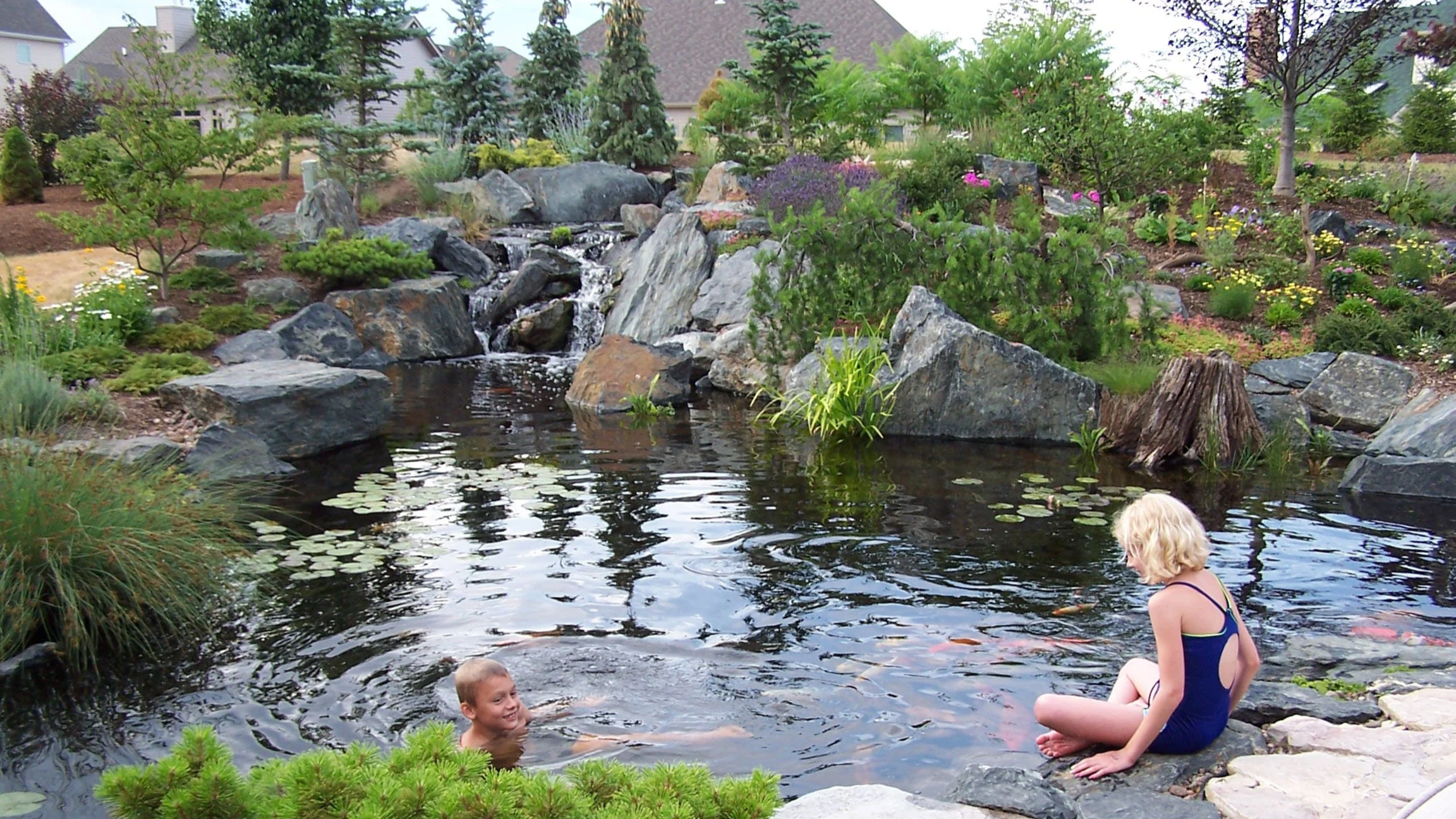 Two children, a boy and a girl, enjoying swimming and sitting by a pond with rocks, surrounded by lush greenery and trees in a backyard garden.