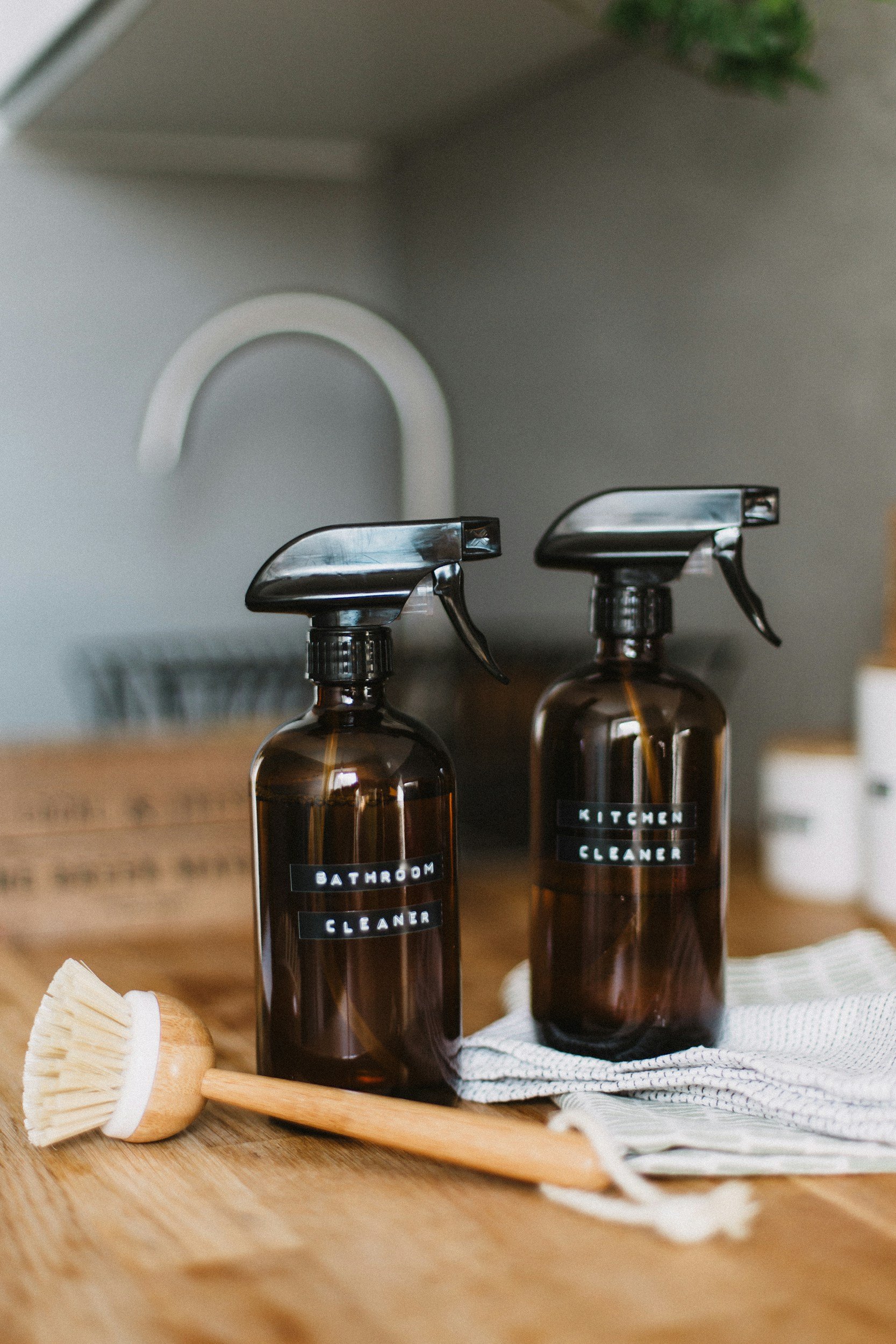 Brown spray bottles labeled "Bathroom Cleaner" and "Kitchen Cleaner" with a wooden scrub brush and cloth on a wooden countertop.