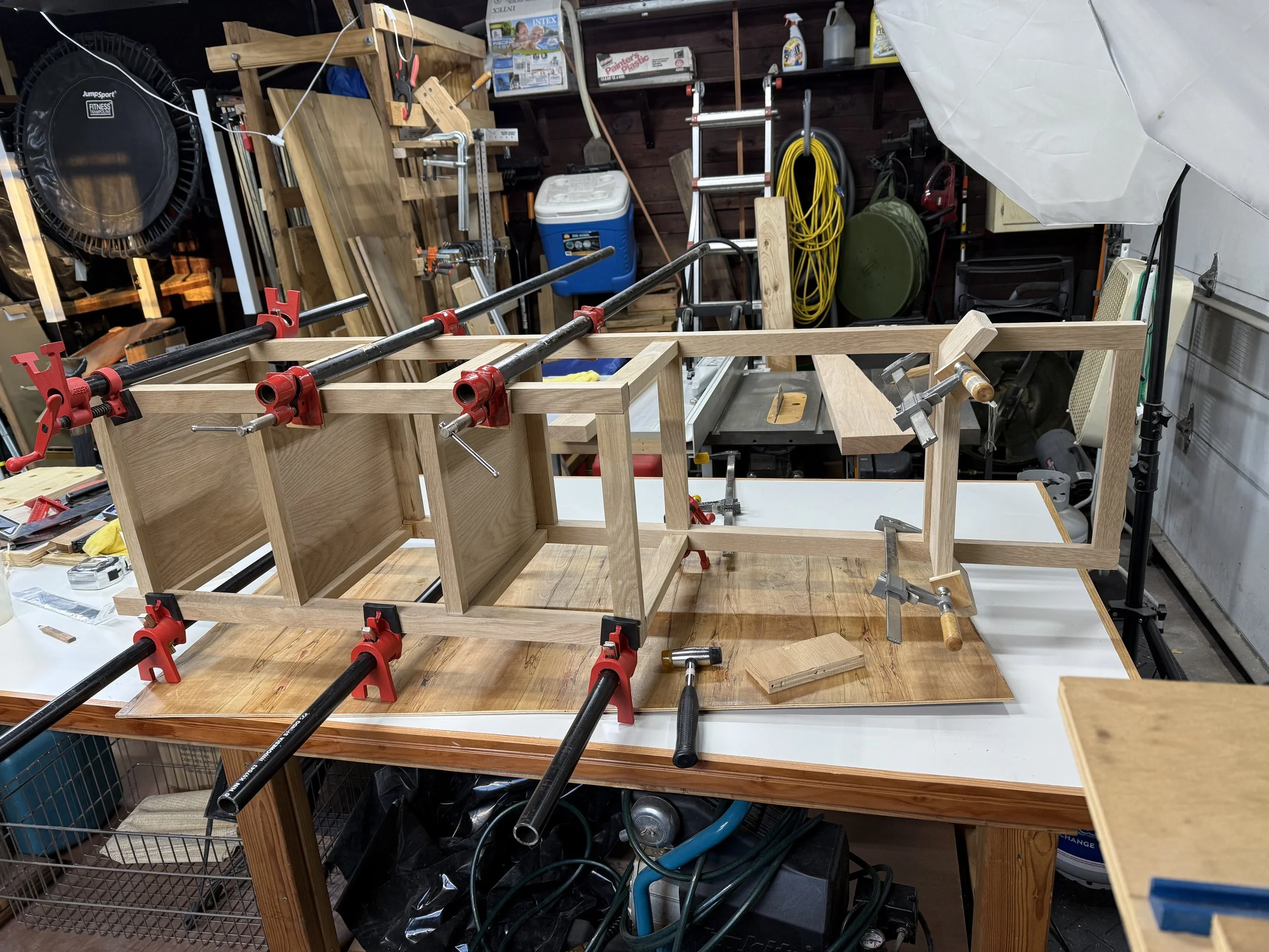 A woodworking project in progress on a workbench, with wooden panels clamped in place for assembly, surrounded by various tools and supplies in a workshop.