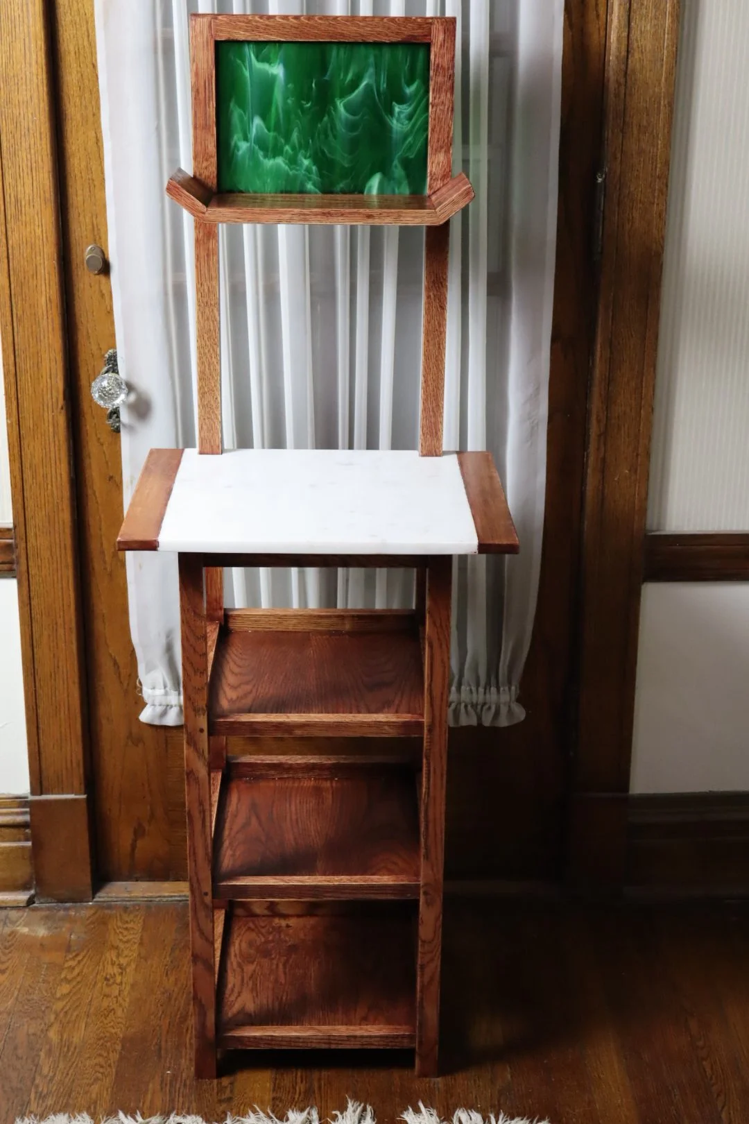 A wooden high chair with a white seat and a green and black marbled backrest, positioned against a wall with white curtains and wooden trim.
