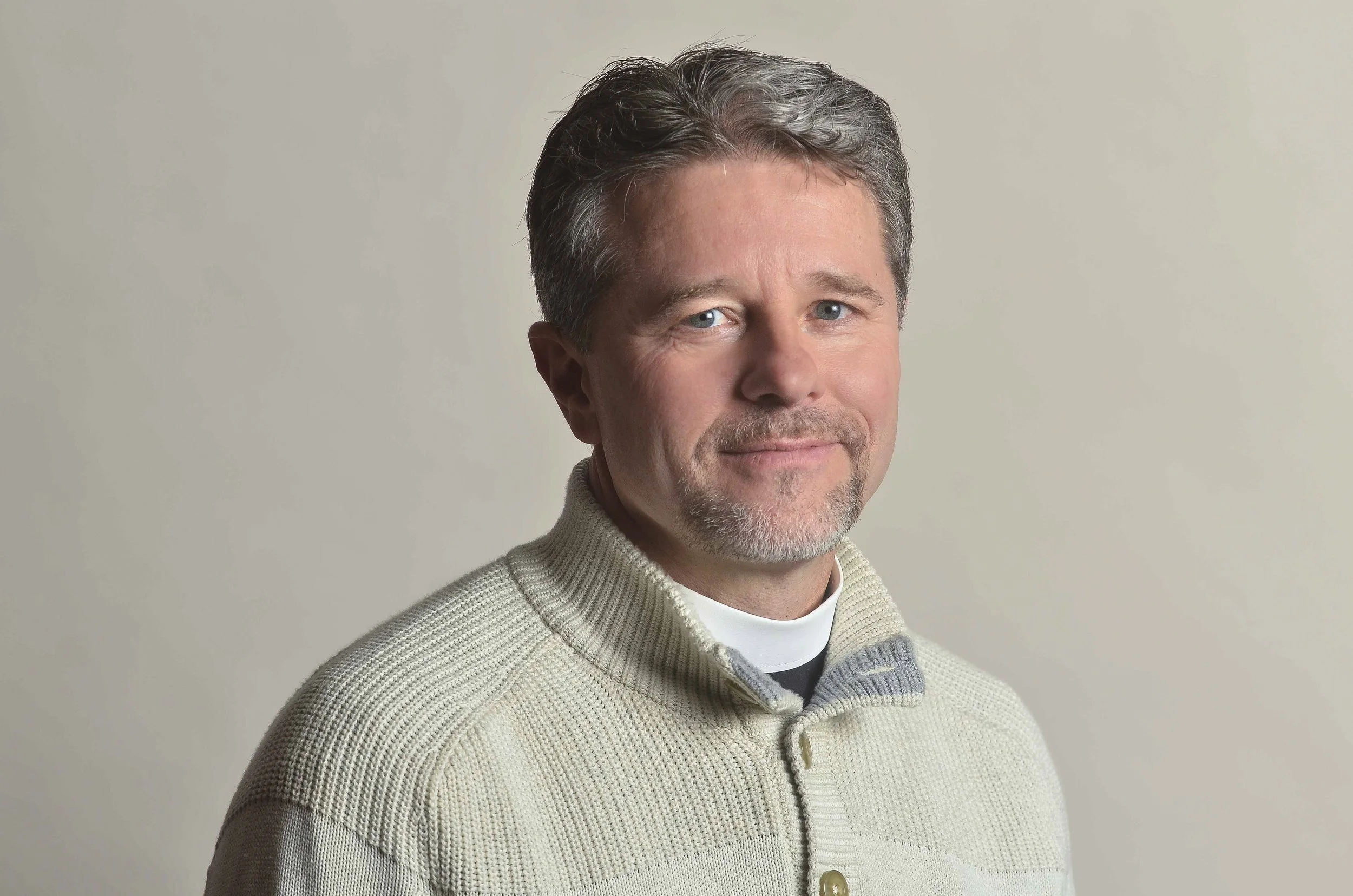 Portrait of the Reverend Matthew McDermott with short, dark and gray hair, light blue eyes, and light skin, wearing a cream-colored knit sweater and a white shirt underneath, smiling gently against a plain light background.