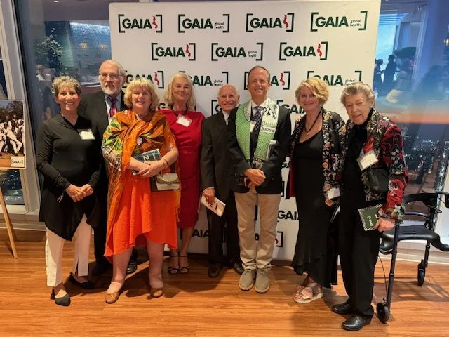 Group of eight diverse people standing together at a Gaia event, posing for a photo in front of a Gaia banner, with city skyline visible through large windows behind them.