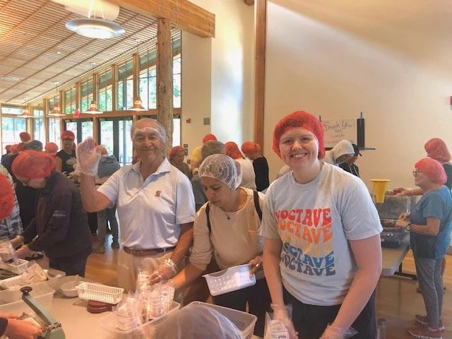 People smiling and working at a food preparation event for Rise Against Hunger, wearing hairnets and gloves, in a large, well-lit room with wood beams.
