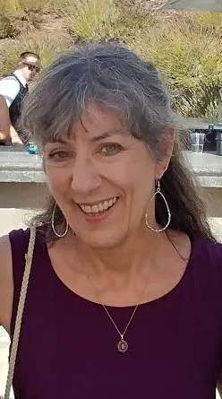 Our Sunday School teacher and ukelele player, Michel Hardbarger, smiling woman with gray hair wearing earrings and a necklace in an outdoor setting.