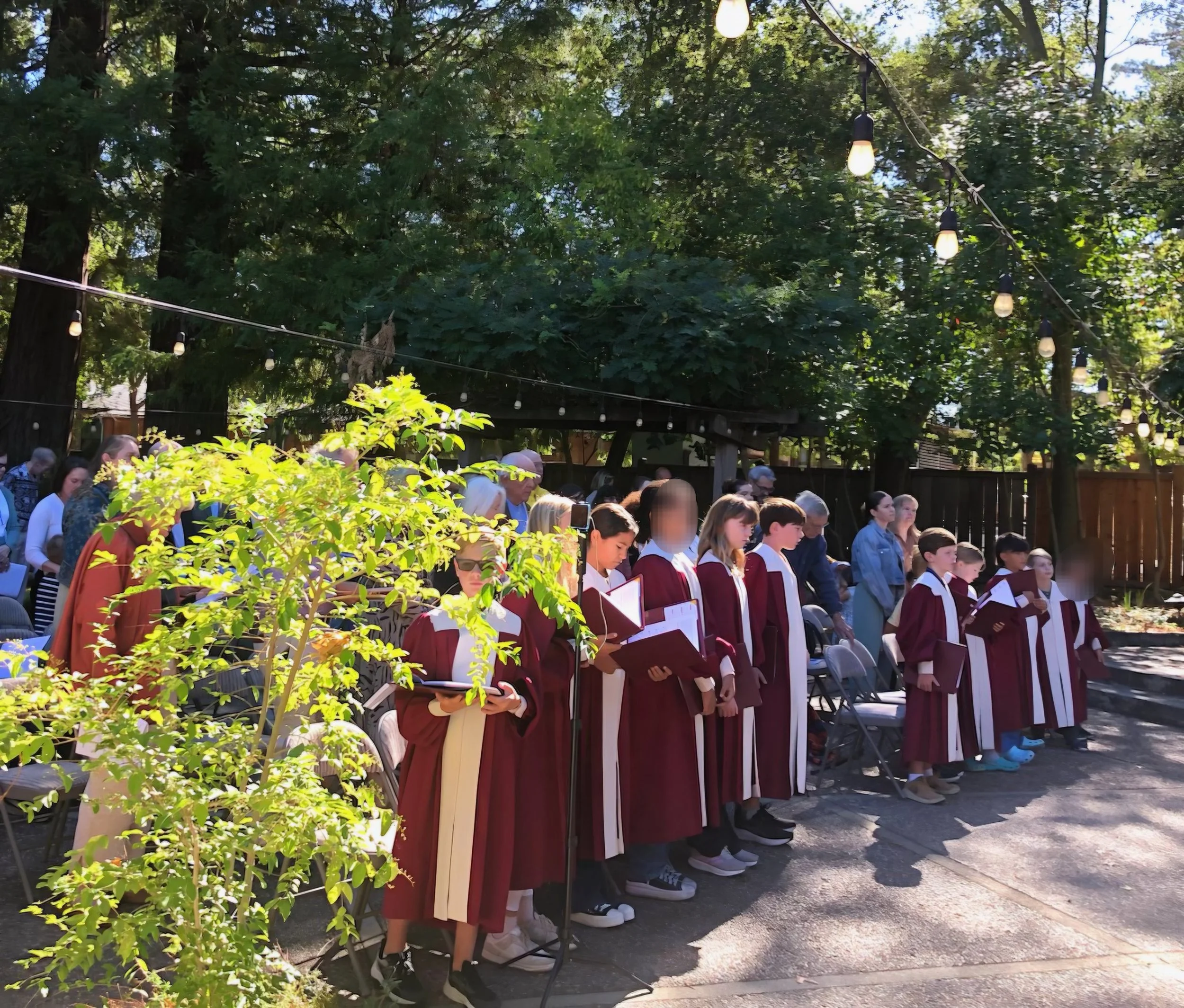 Christ Church children's choir, at our outdoor Blessing of Animals service held in October. Trees and string lights are overhead, and there are chairs and a wooden fence in the background.