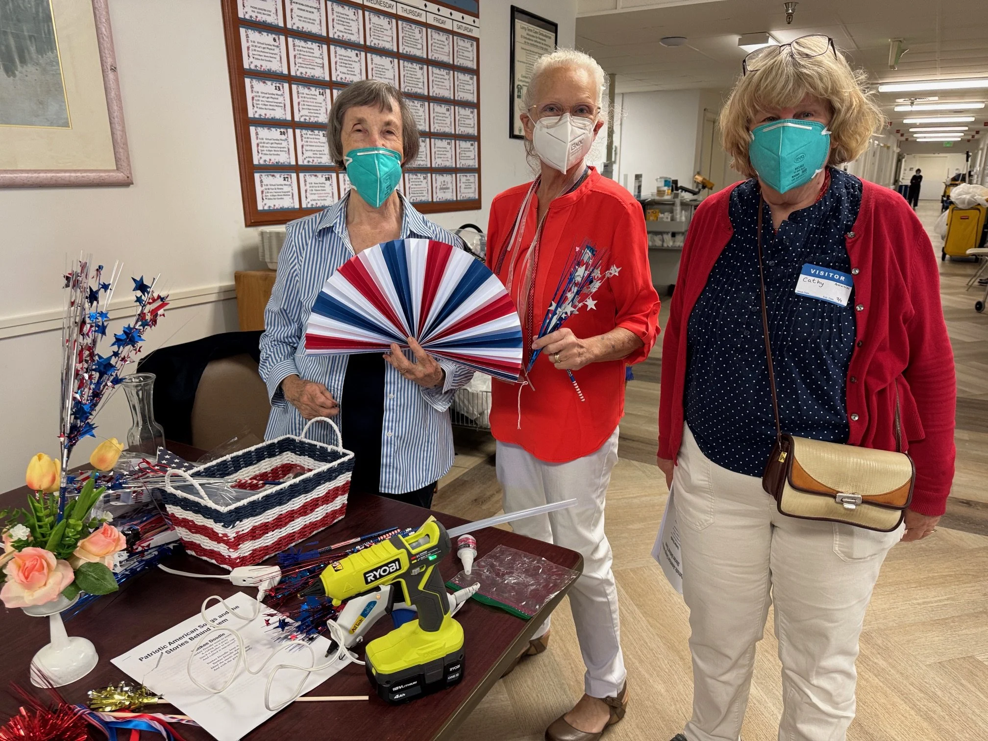 Three women wearing masks at Burlingame Skilled Nursing, where they led activities for SpiritCare Ministry, standing behind a table decorated with patriotic items, including a large paper fan, ribbons, and flowers for July 4th.