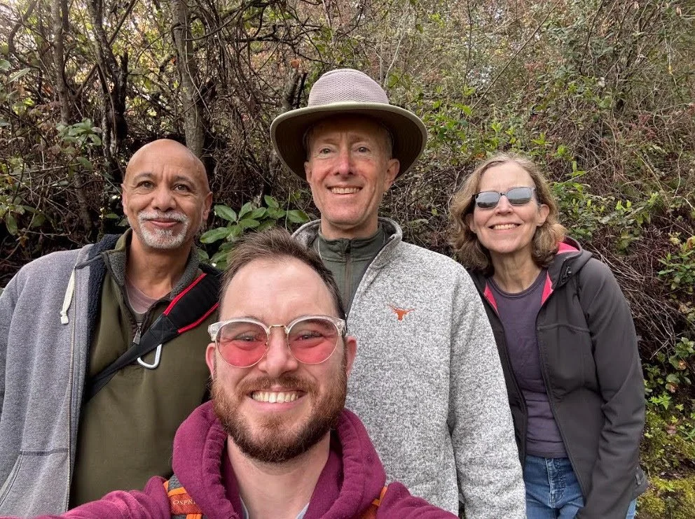 Four people hiking outdoors in a wooded area, smiling for a selfie. Two men and two women, dressed in casual outdoor attire.