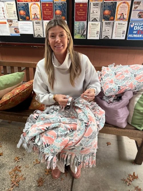 A woman sitting on a wooden bench with colorful pillows, holding a blanket with a floral pattern, in front of a display of various posters or flyers on a wall.