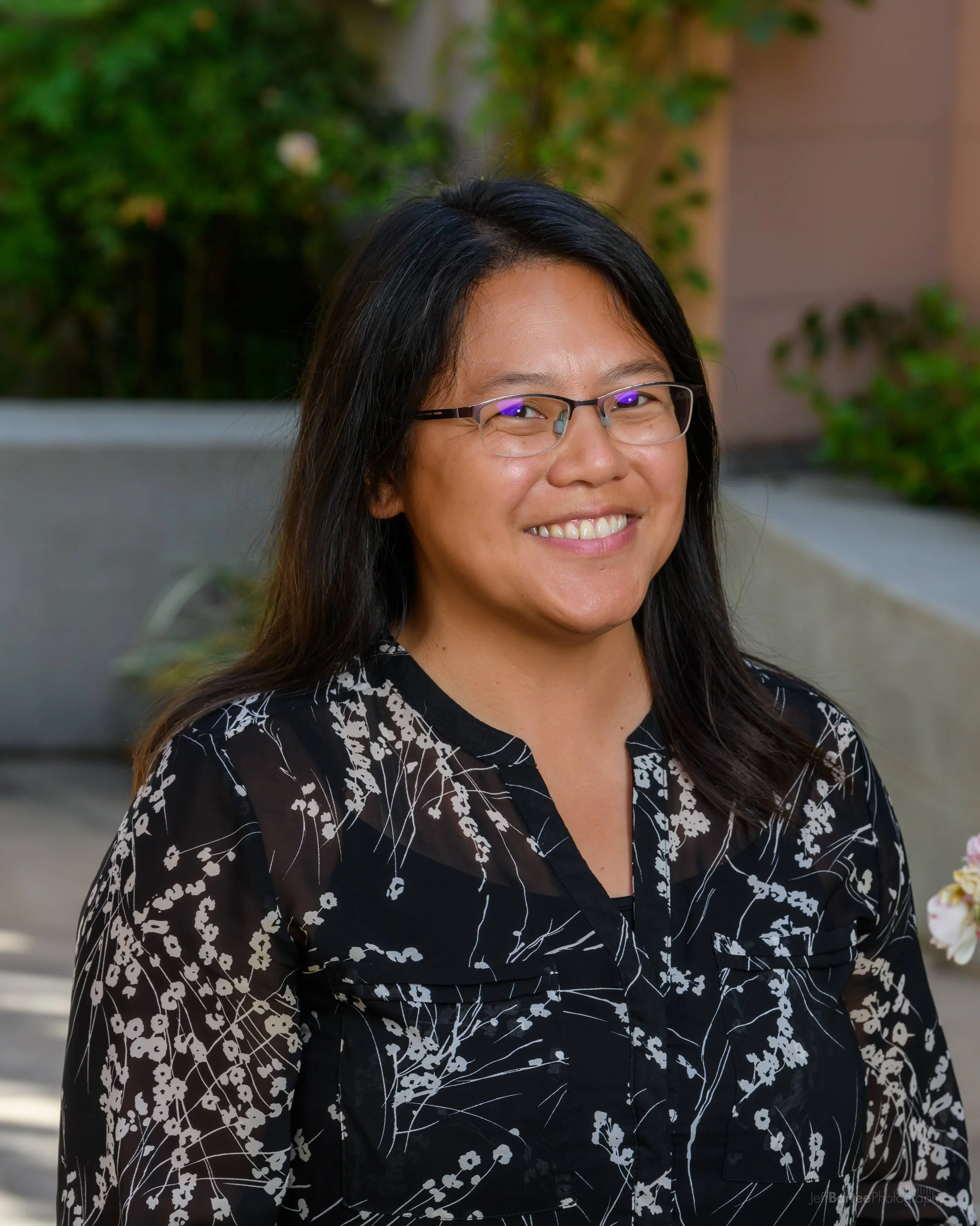 Our program director, Jocelyn Sideco, wearing glasses and a black blouse with white floral patterns, smiling outdoors with greenery in the background.
