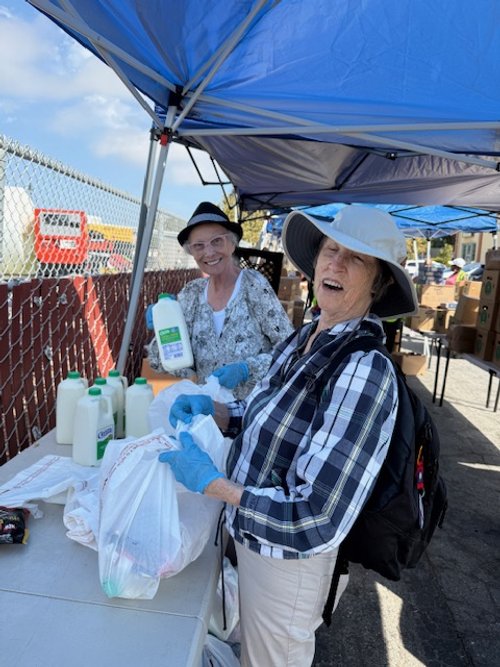 Two women wearing hats and gloves standing under a blue canopy in East Palo Alto handing out food and supplies.