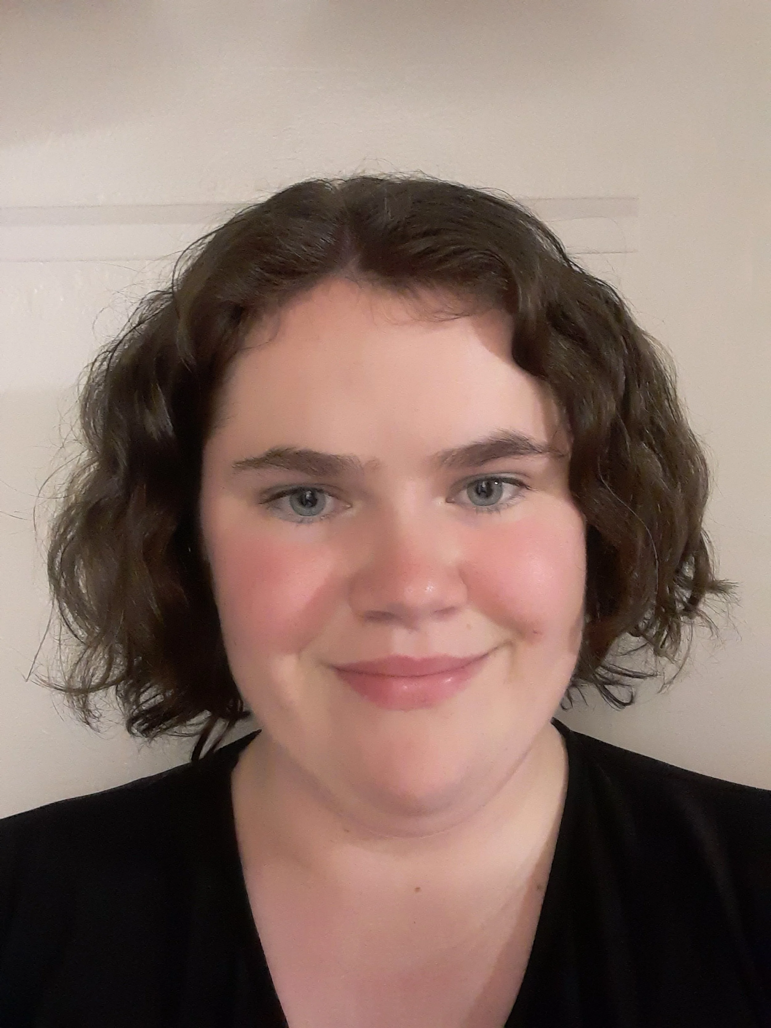 Our children's choir director, Katy Darrow, woman with short, curly brown hair and blue eyes smiling at the camera against a plain off-white wall.