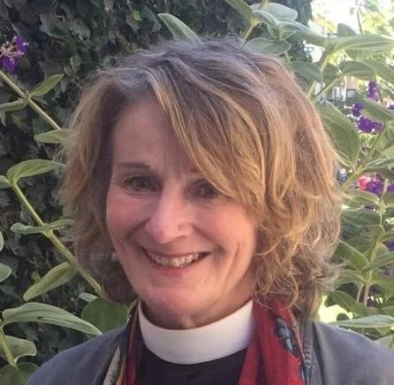 The Reverend Lauren McCombs, a deacon, with curly blonde hair smiling outdoors with green foliage and purple flowers in the background.