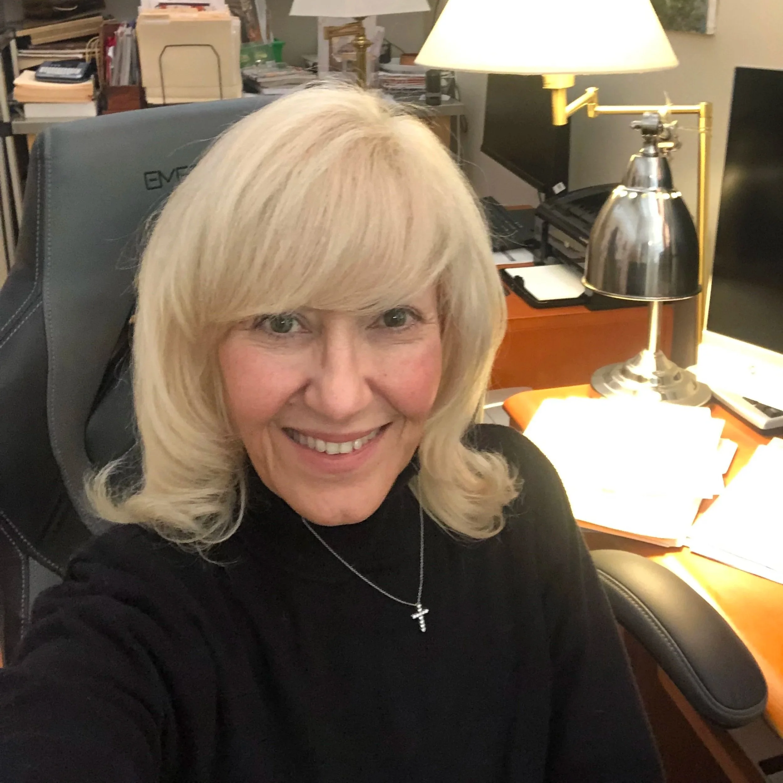 A smiling Chantal, our parish administrator, with blonde hair in her office, with a desk, lamp, and computer in the background.
