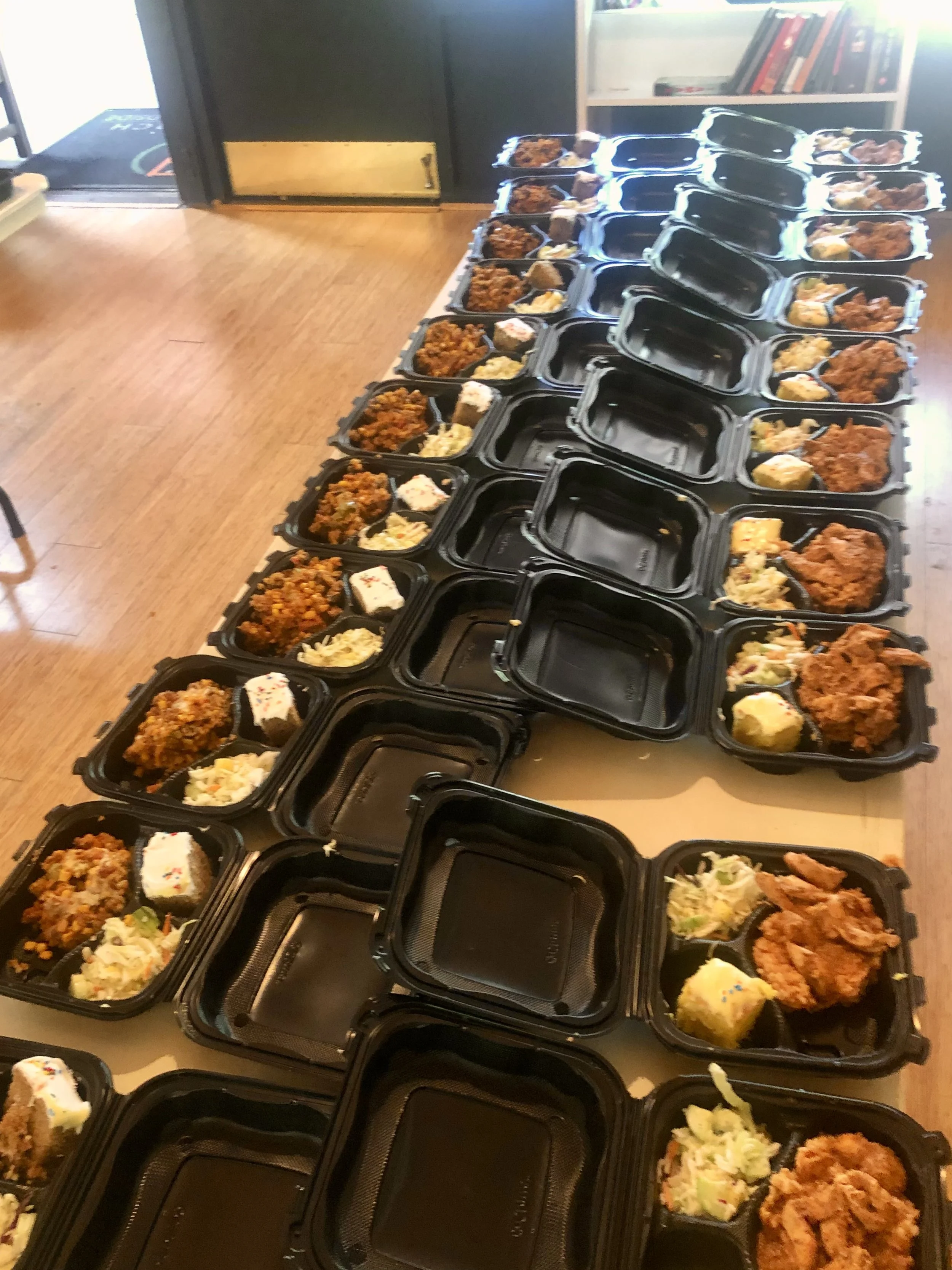 Multiple black meal containers with fried chicken, potato salad, and cake on a table.