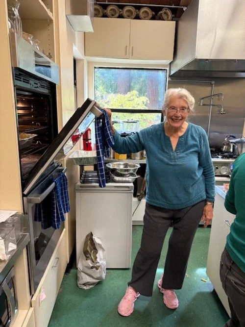 Smiling elderly woman, Ruth Ann Wrucke, in a kitchen holding a dish towel, wearing a blue sweater and gray pants.