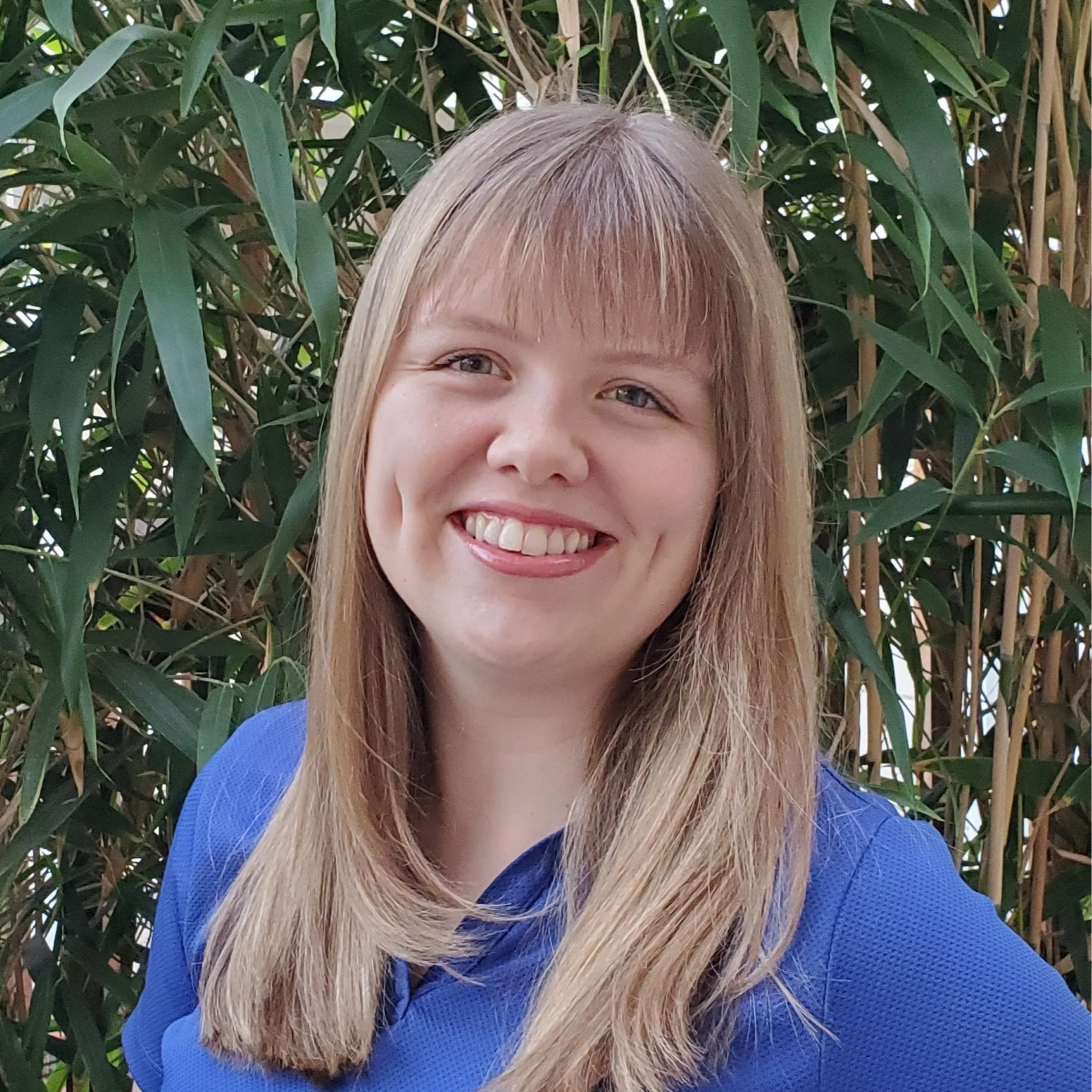 Smiling young woman, our music director, Lauren Wittine, with long blonde hair wearing a blue shirt standing in front of green plants.