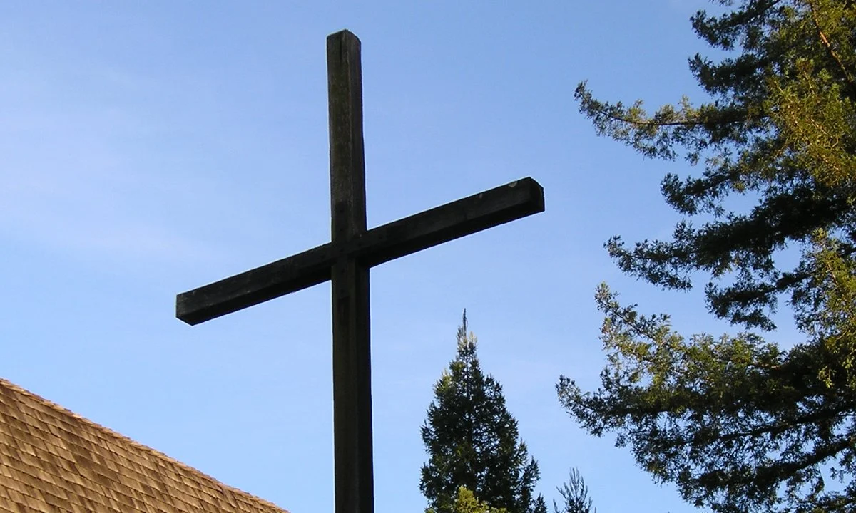 Wooden cross on a church roof with trees and a blue sky in the background.