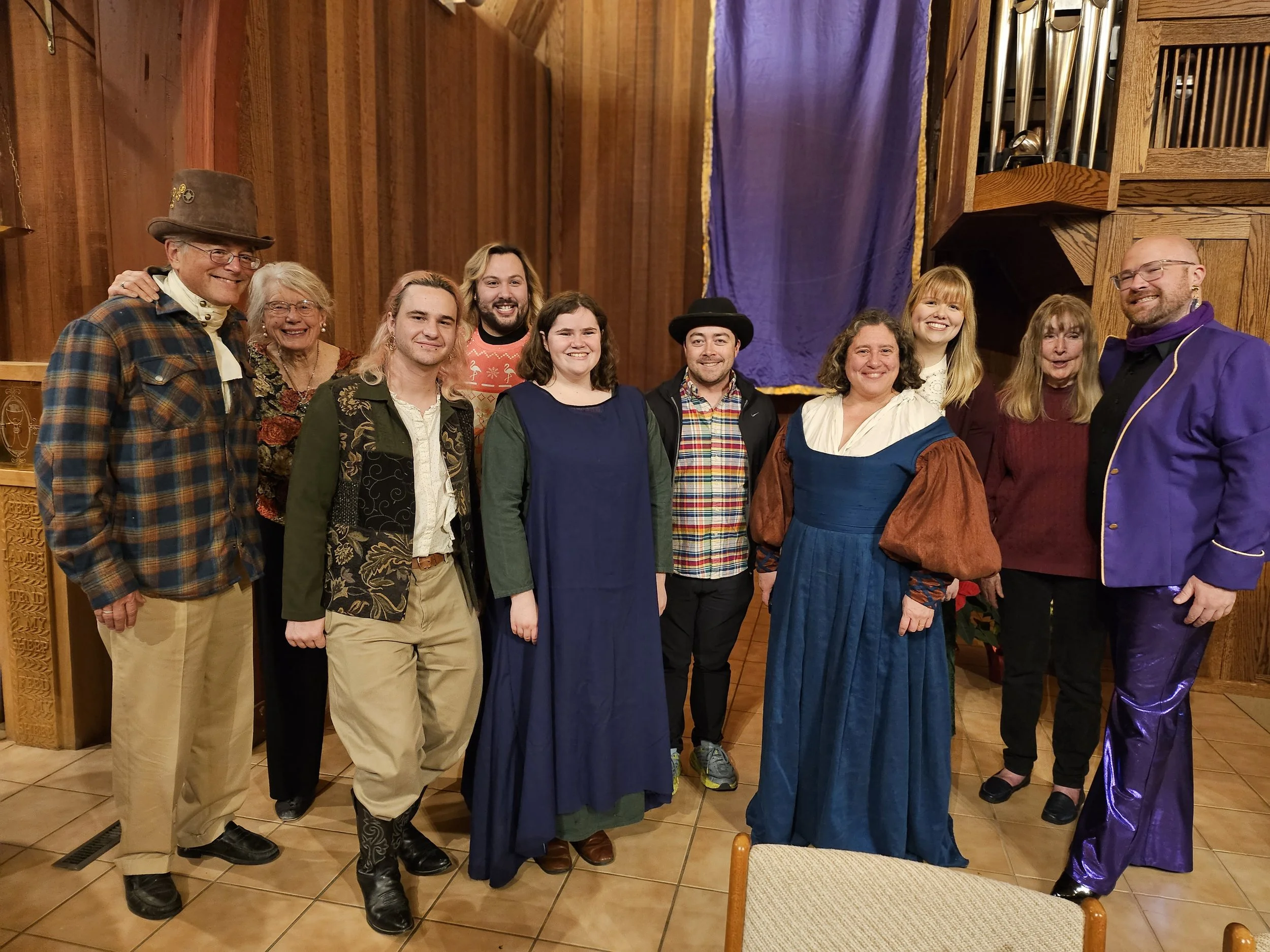 Christ Church Choir Members dressed in historical or theatrical costumes, standing indoors in front of wooden walls and a purple curtain.
